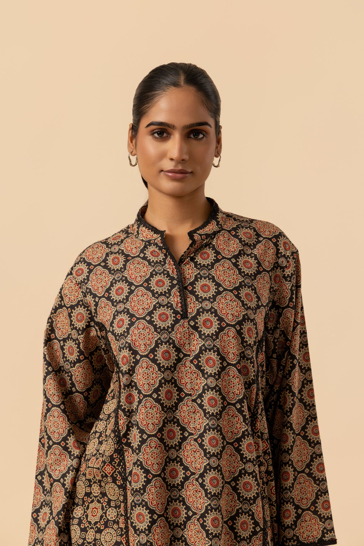 A woman wearing a long-sleeve ajrakh co-ord set with an intricate geometric and floral pattern in brown, rust, and black, set against a solid beige background.