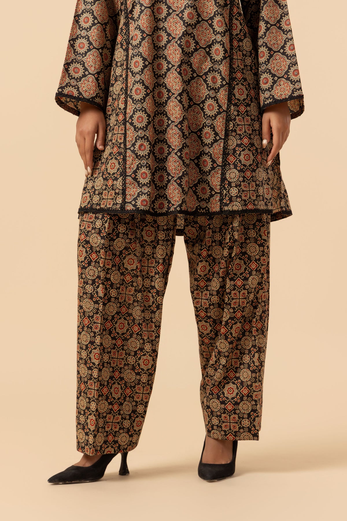 A model is shown from the waist down wearing a matching two-piece ajrakh co-ord set with a brown, black, and beige geometric pattern, paired with black pointed-toe heels, against a solid beige background.