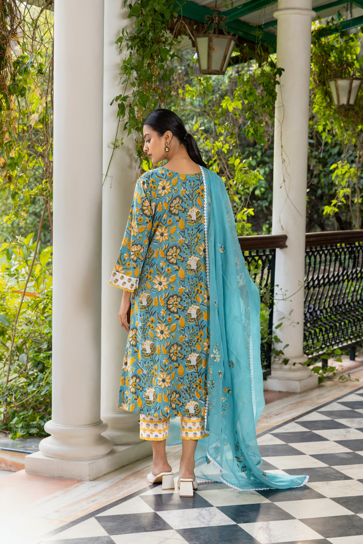 Back view of a person wears a vibrant turquoise and yellow handblock printed kurta set, standing gracefully on a checkered floor amidst lush greenery.
