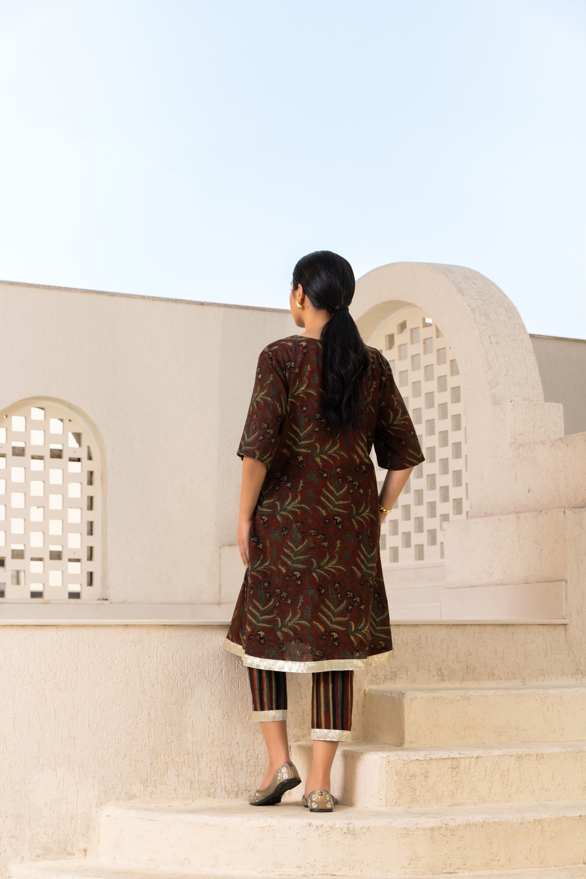 Back View of a Model wearing a brown printed kurta set with intricate patterns, paired with striped trousers, styled in a serene outdoor setting
