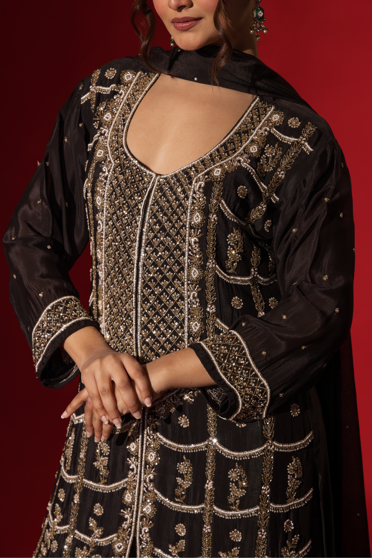 Closeup view of a woman's hand wearing elegant black embroidered kurta set with intricate gold embroidery against a rich red backdrop.