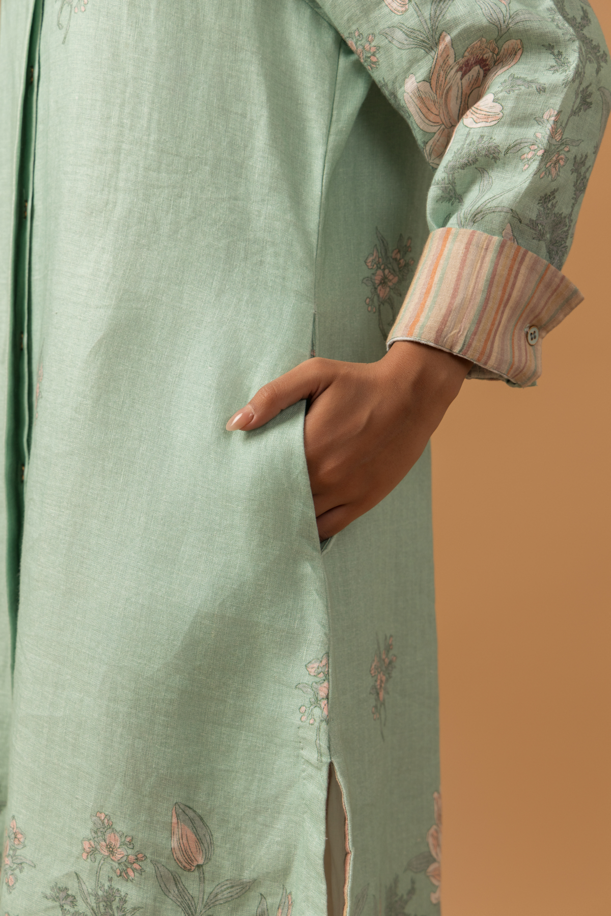 A person wearing a light green floral dress with a striped cuff has their right hand in a pocket.