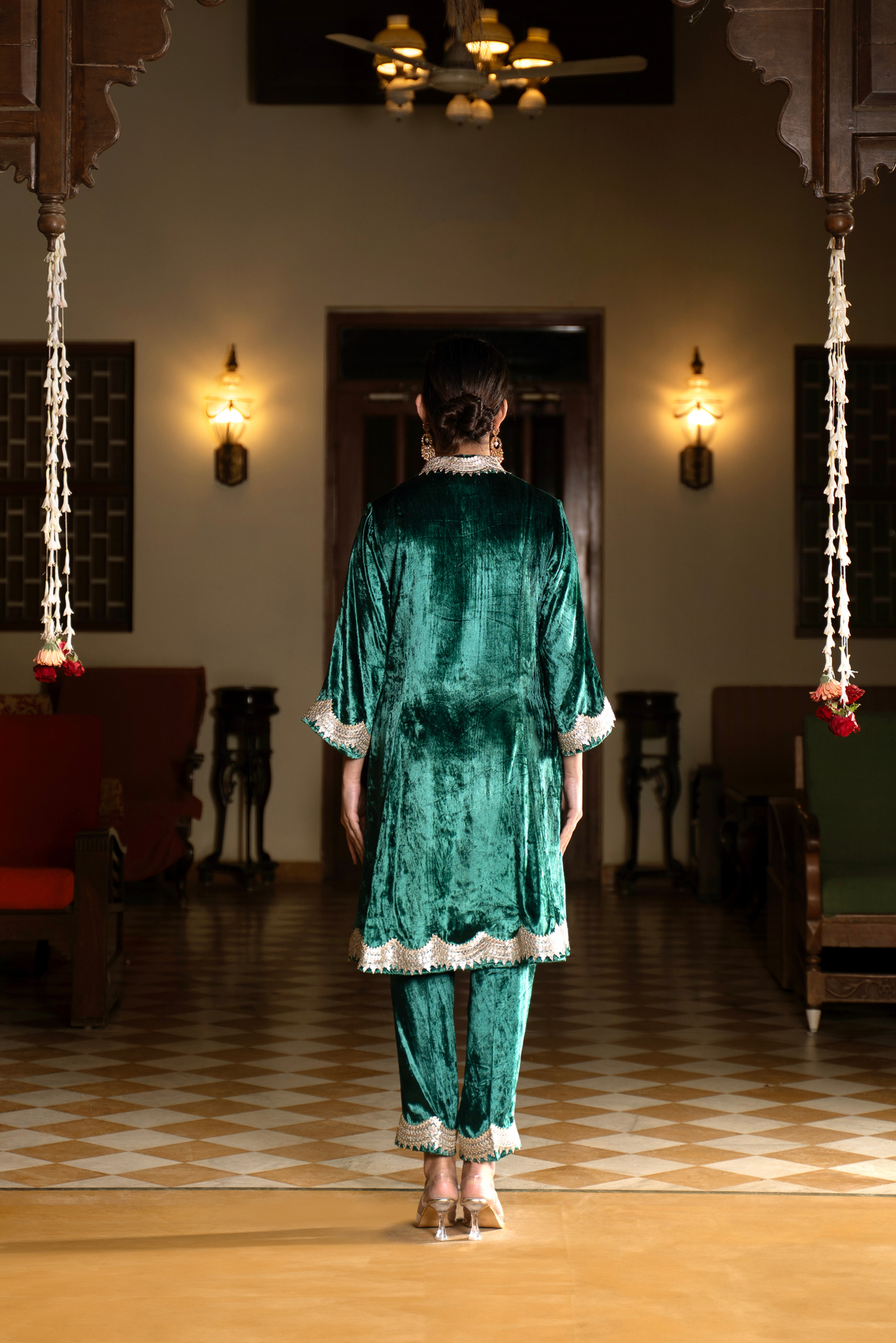 Back view of a woman in a green velvet co-ord set traditional outfit stands in a warmly lit room with decorative lighting and patterned flooring.