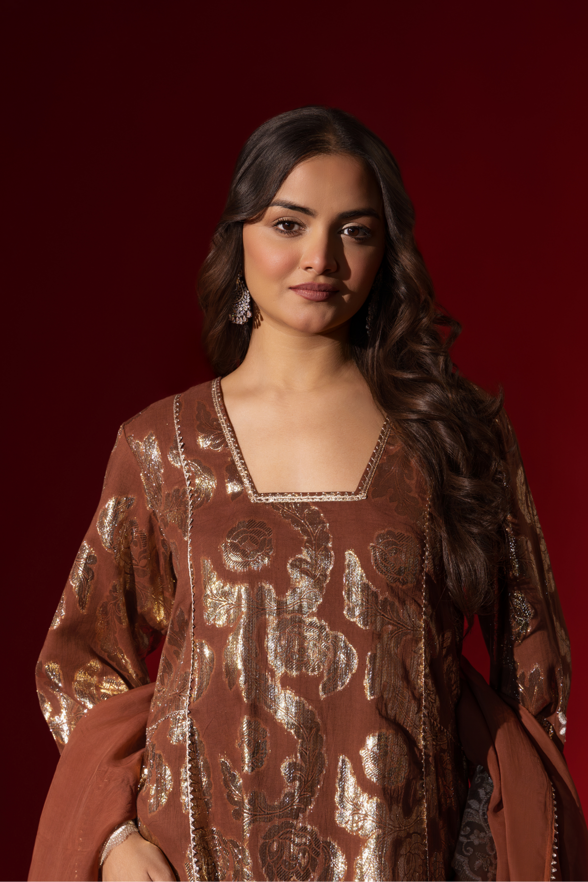 Closeup view of a model poses in a brown ethnic shimmer kurta set outfit with intricate white embroidery against a vibrant red backdrop.
