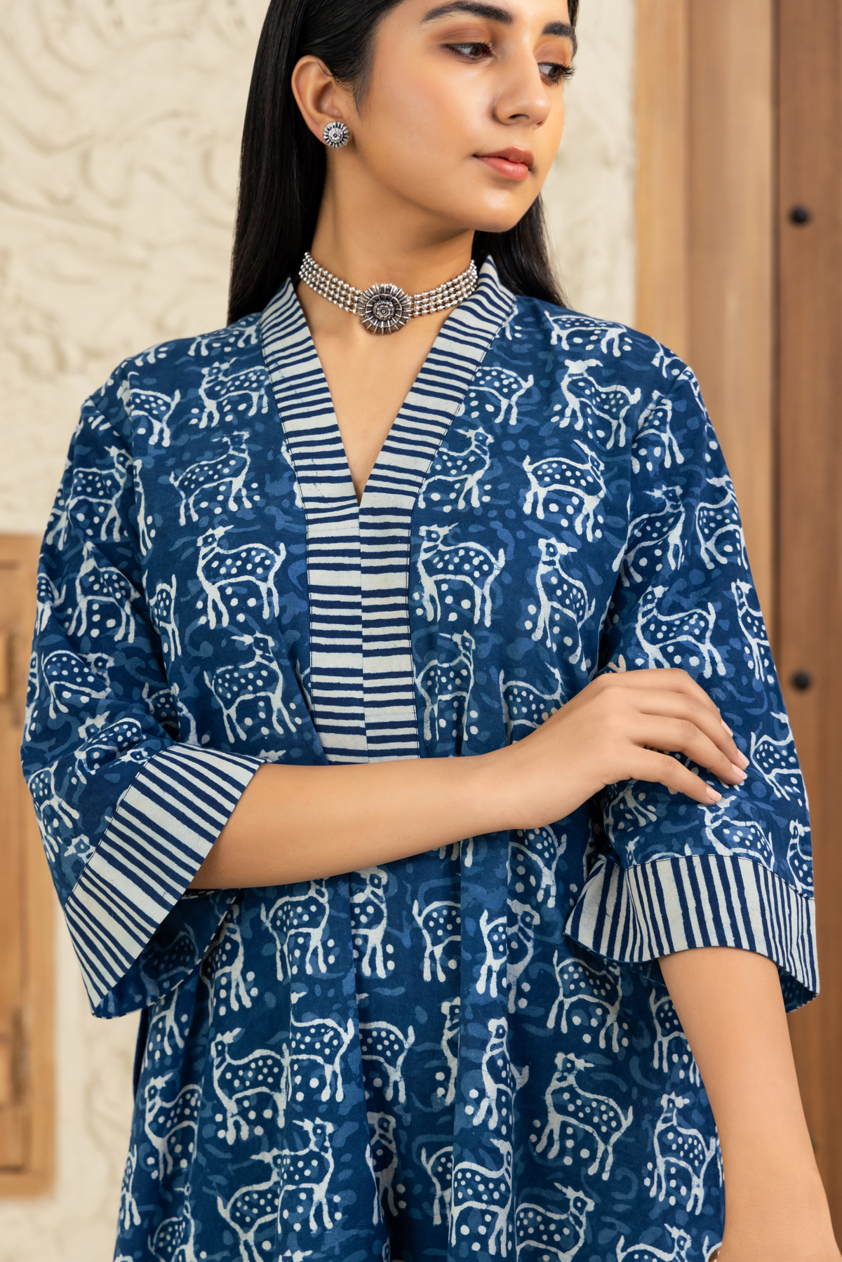 closeup view of a model dressed in a blue and white deer printed co-ord set with striped detailing, standing against a wooden and textured beige background.