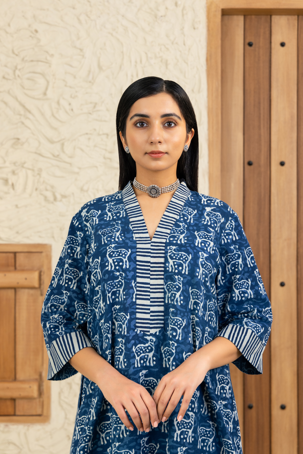 Closeup view of a woman wearing a blue and white deer printed co-ord set with striped detailing, standing against a wooden and textured beige background.