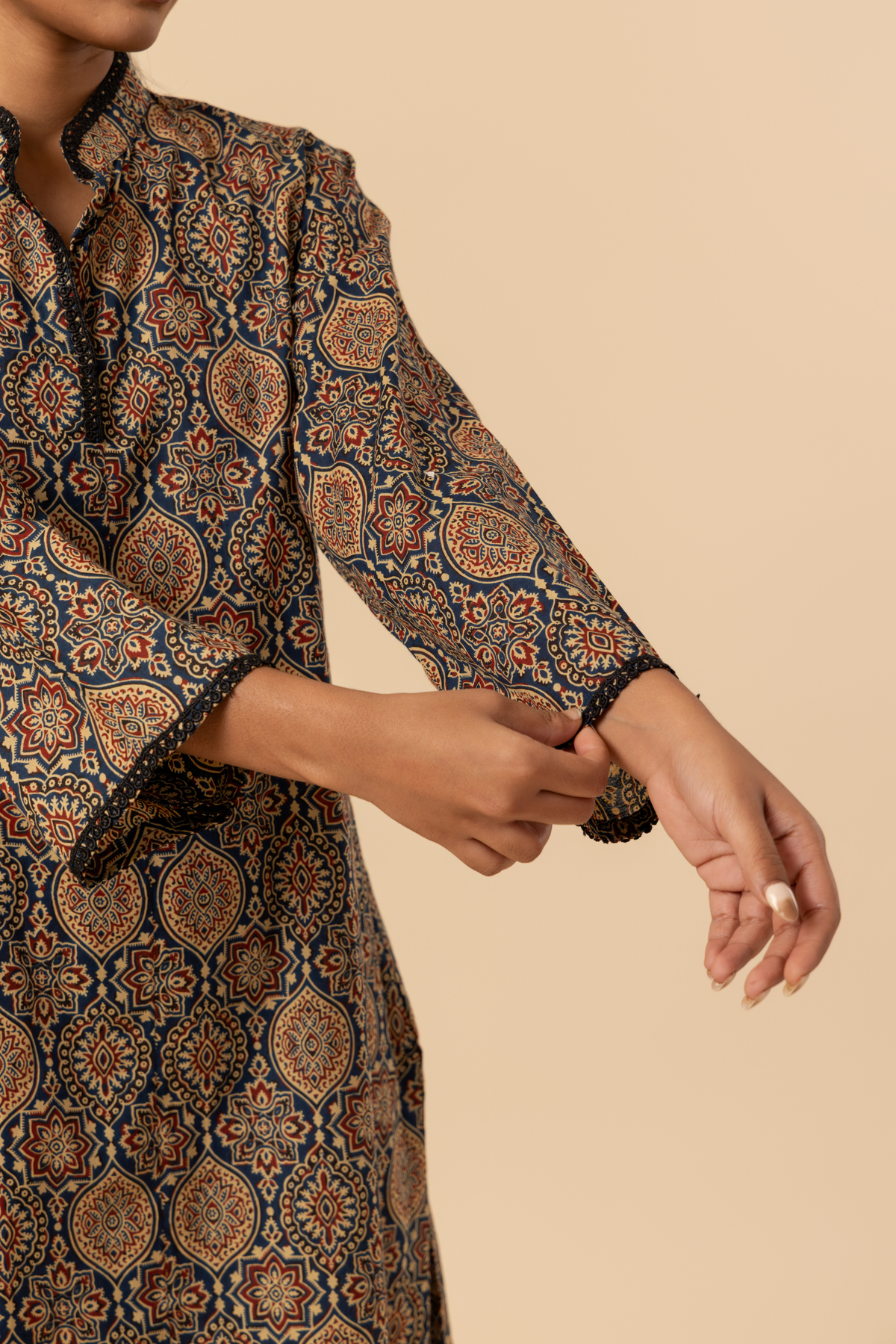 Closeup view of a model's hand stands in a patterned ajrakh print co-ord set which has brown kurta and matching loose pants against a soft beige background, showcasing traditional ethnic wear.