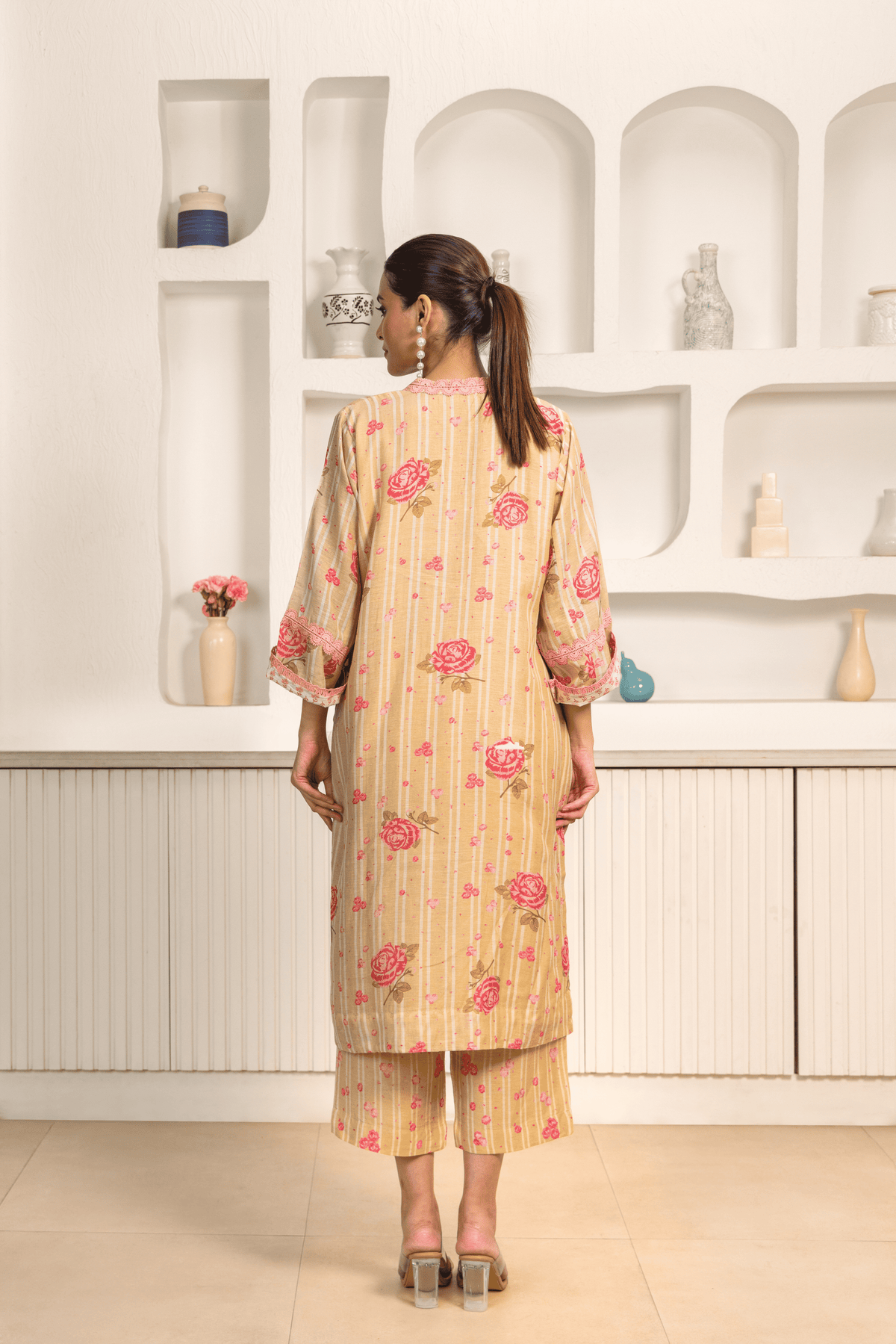 Back view of model wearing a golden floral printed long tunic featuring pink flowers with matching cropped pants, paired with clear heels in a minimalist interior setting.