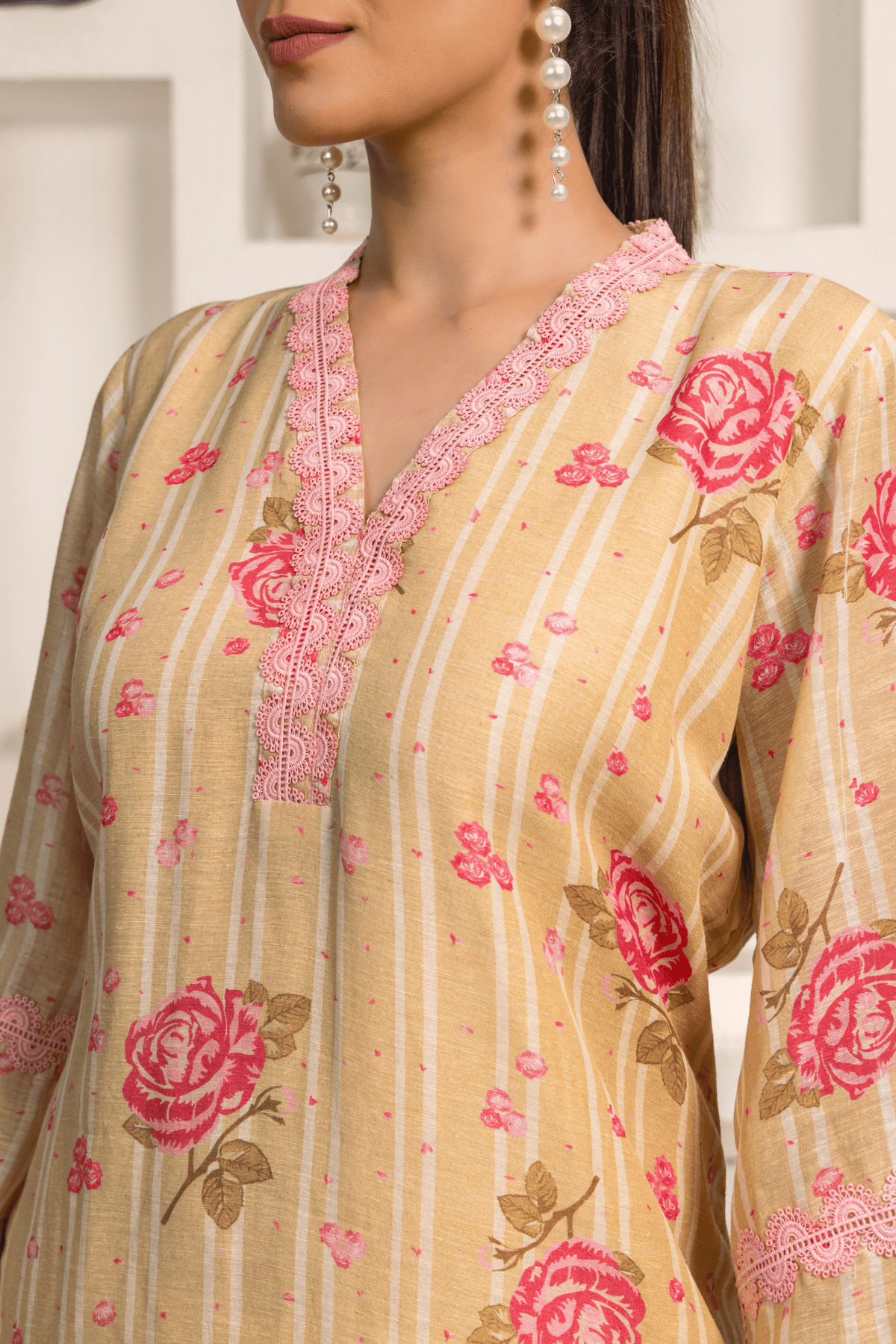 Close up view of model wearing a golden floral printed long tunic featuring pink flowers with matching cropped pants, paired with clear heels in a minimalist interior setting.