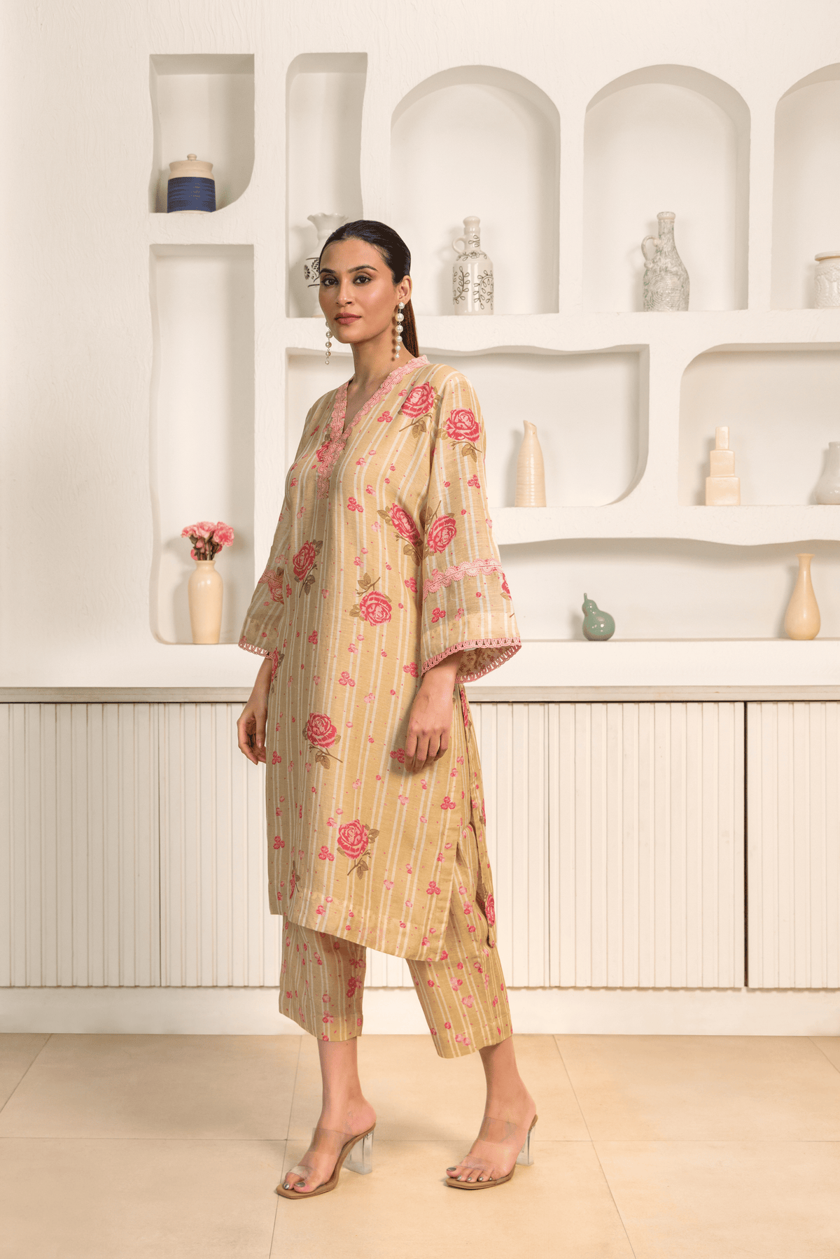 A model wearing a golden floral printed long tunic featuring pink flowers with matching cropped pants, paired with clear heels in a minimalist interior setting.