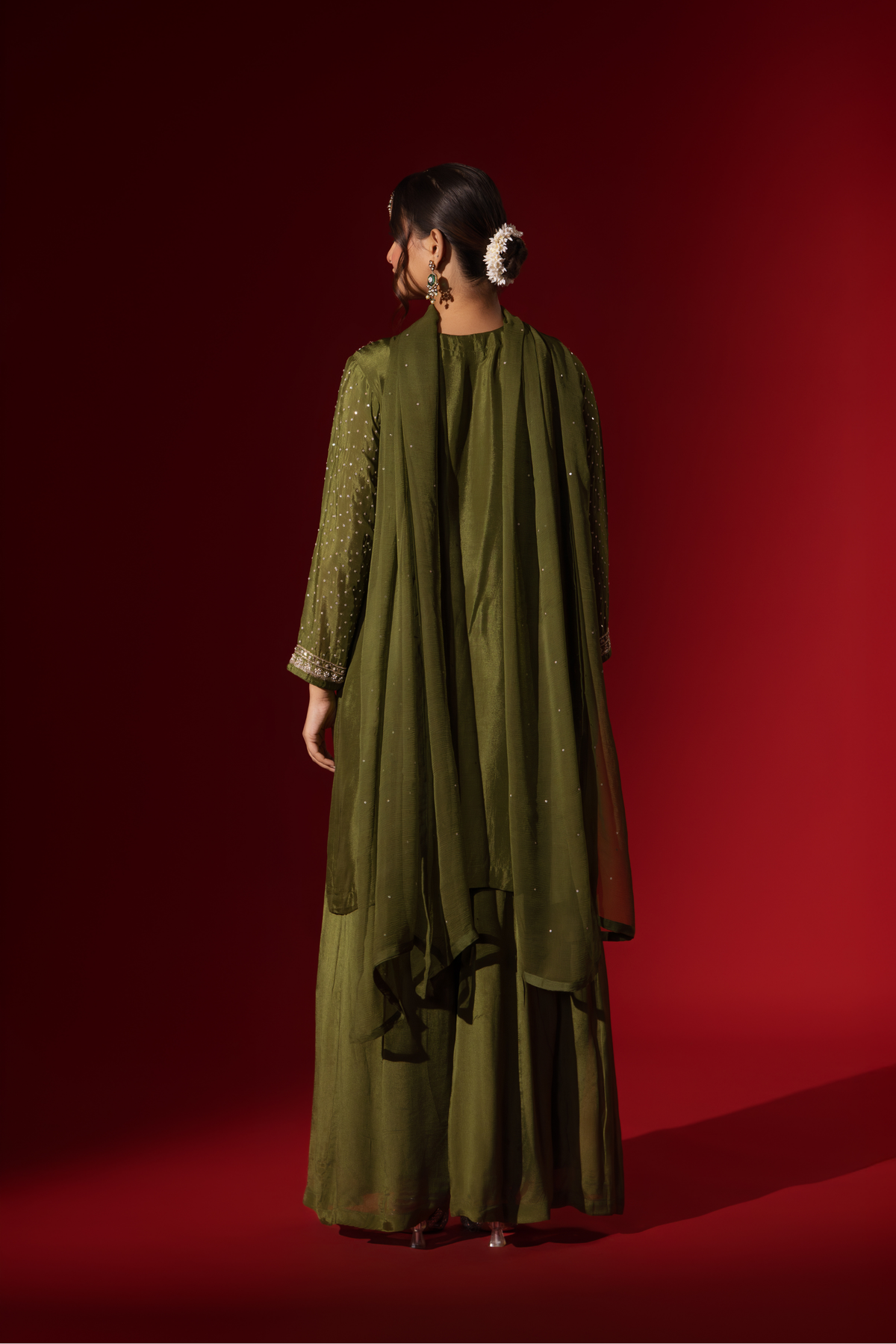 Back view of a woman poses in an elegant olive green embroidered kurta set with intricate floral embroidery, set against a rich red backdrop.
