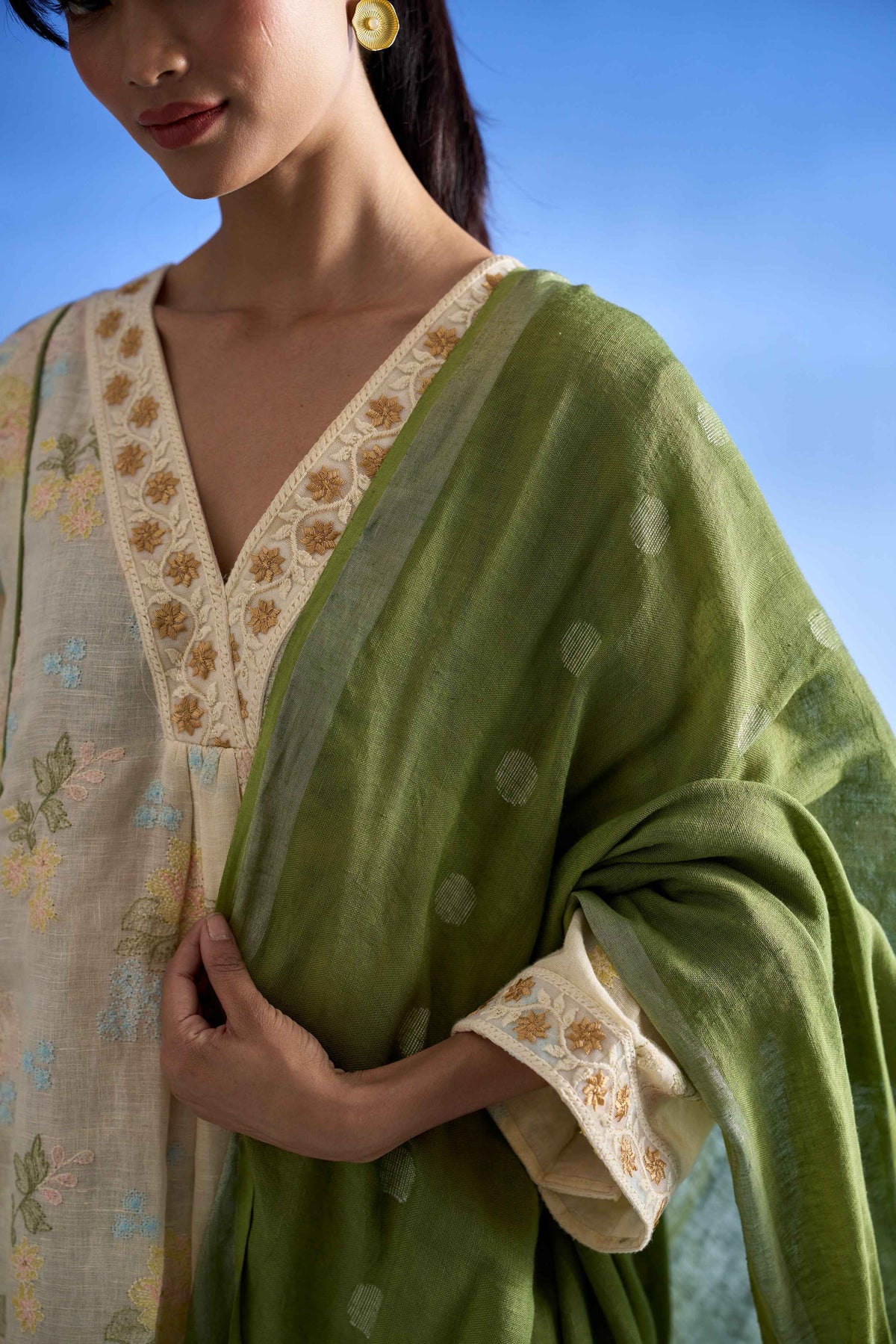 A close-up of a woman wearing a green dupatta draped over a cream-colored embroidered kurta, set against a clear blue sky background.