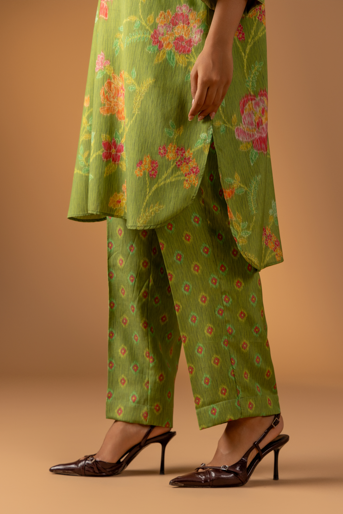 A close-up of a woman wearing a vibrant green outfit with floral patterns and chic brown heels against a soft, warm background.