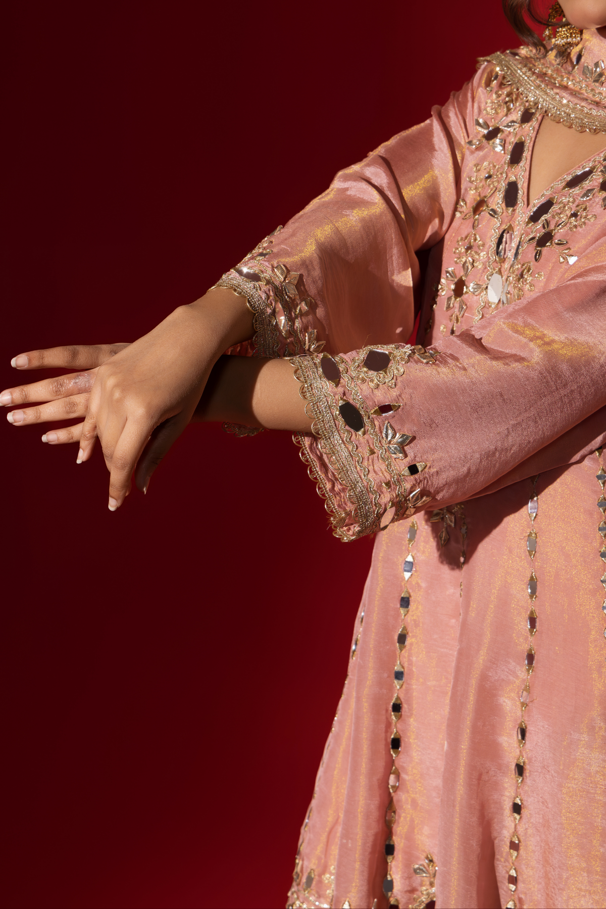 Closeup view of a woman's hand against a red background, wearing an elegant pink embroidered kurta set intricately embellished outfit with a matching shawl.