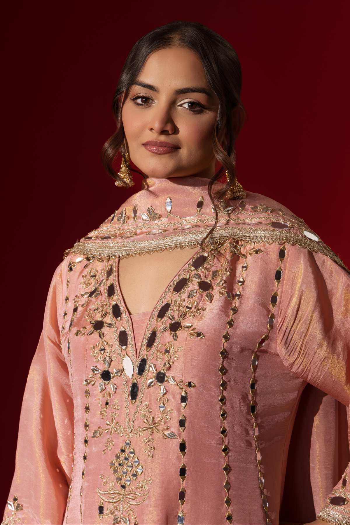 Closeup view of a person stands against a red background, wearing an elegant pink embroidered kurta set intricately embellished outfit with a matching shawl.