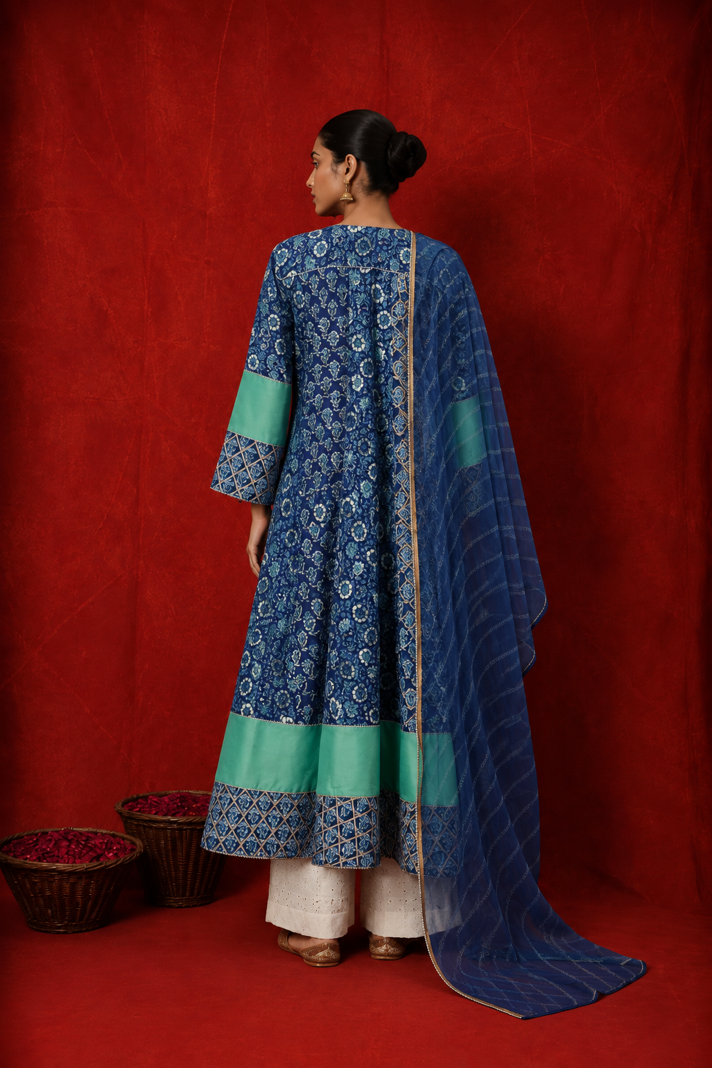 Backside of a woman wearing a blue floral printed kurta with white pants and a matching blue dupatta featuring green borders, posed against a rich red backdrop.