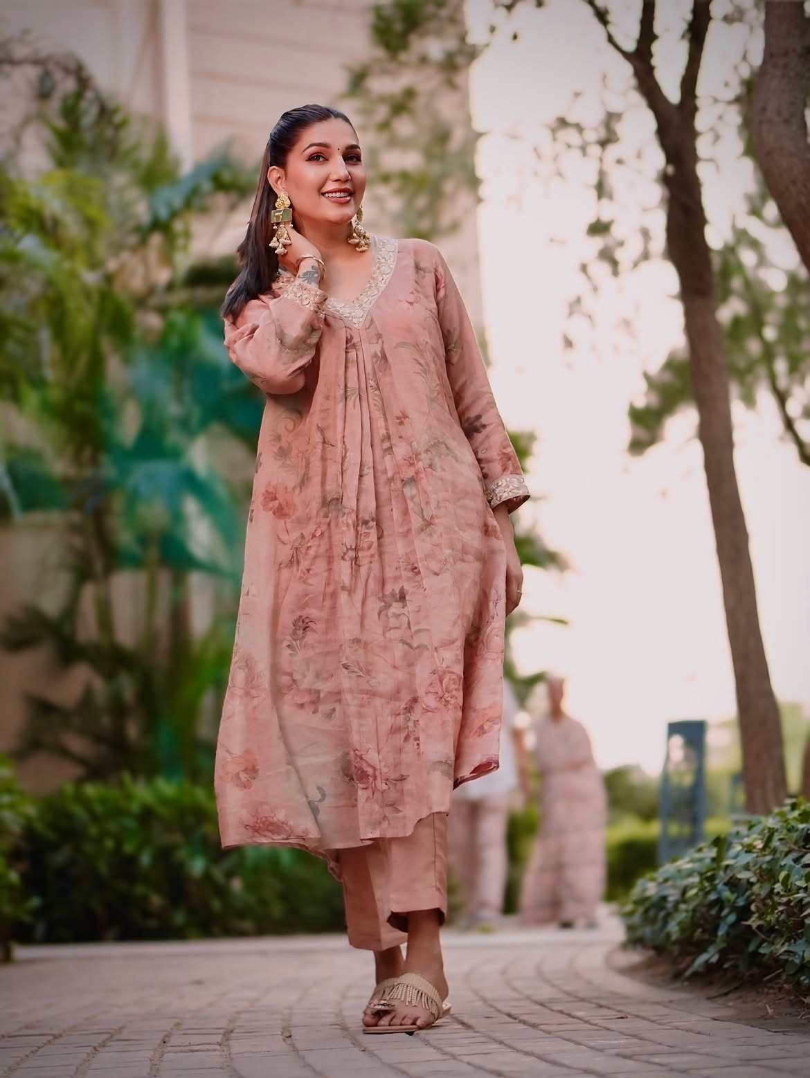 A woman wearing a dusty pink floral printed kurta and matching trousers walks outdoors on a paved path with trees and greenery in the background.