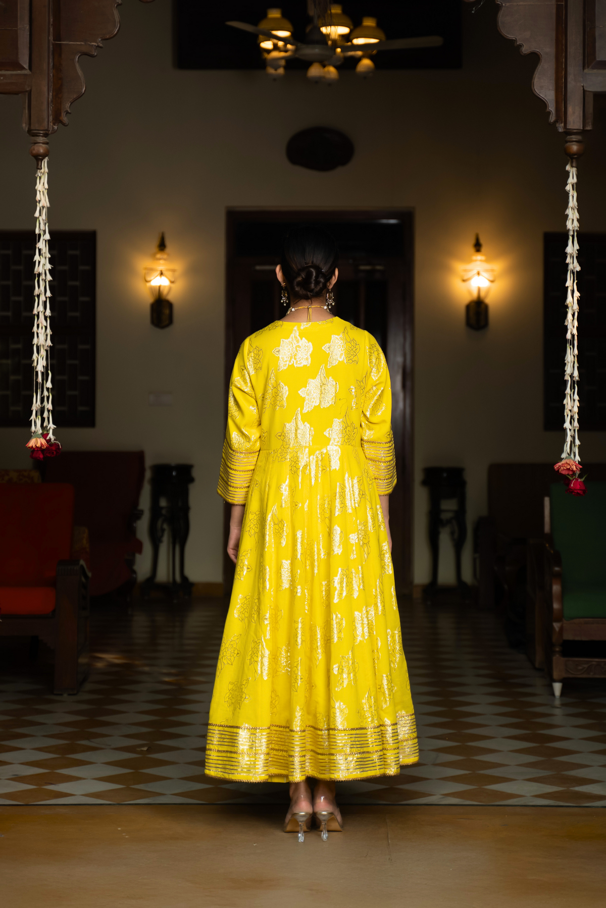 Back view of a woman in a yellow anarkali kurta with intricate gold patterns with matching pants and dupattaa, standing in a beautifully decorated interior.
