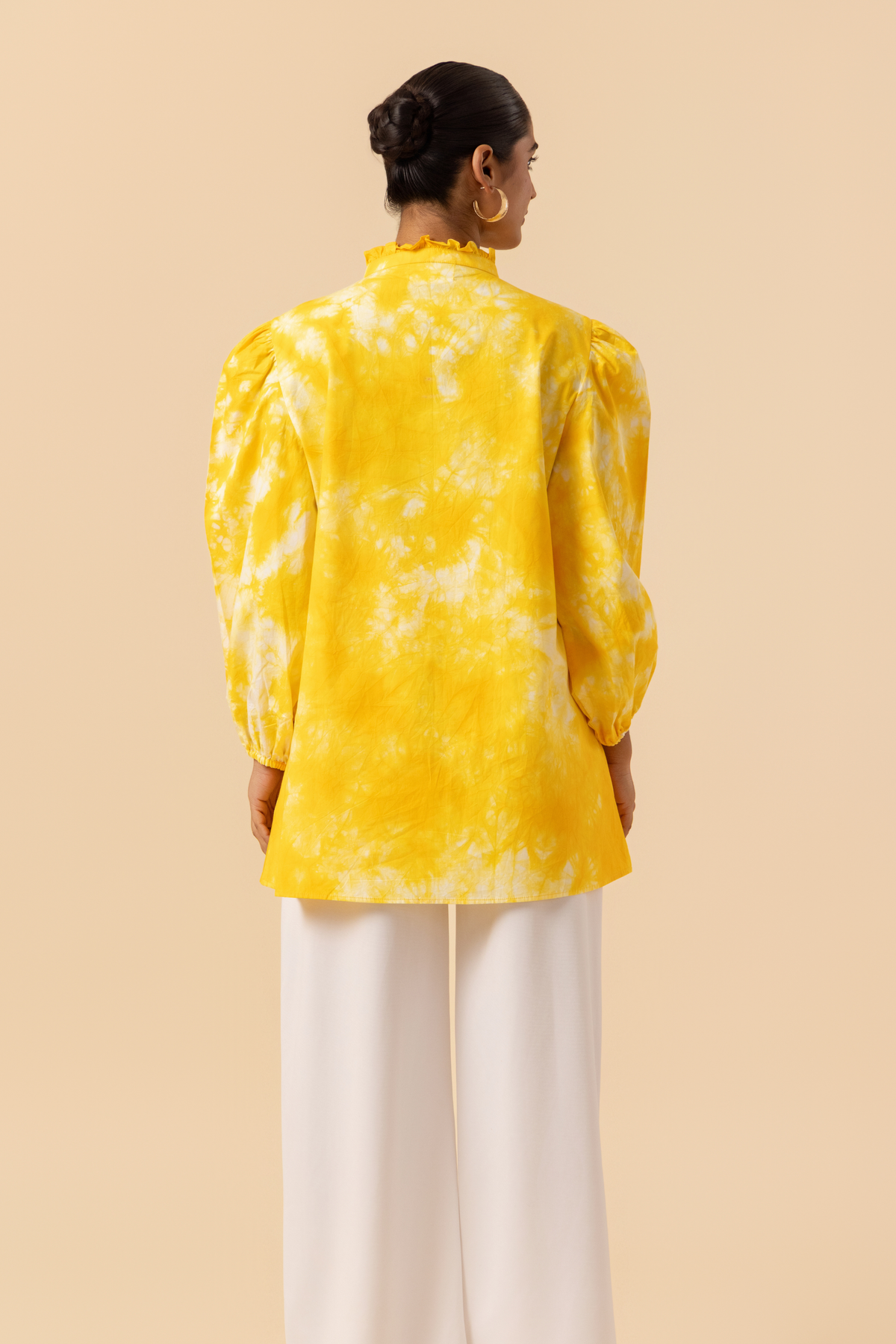 Back view of a woman wears a vibrant yellow tie-dye shirt dress with puffed sleeves, paired with white wide-leg trousers and stylish sandals.