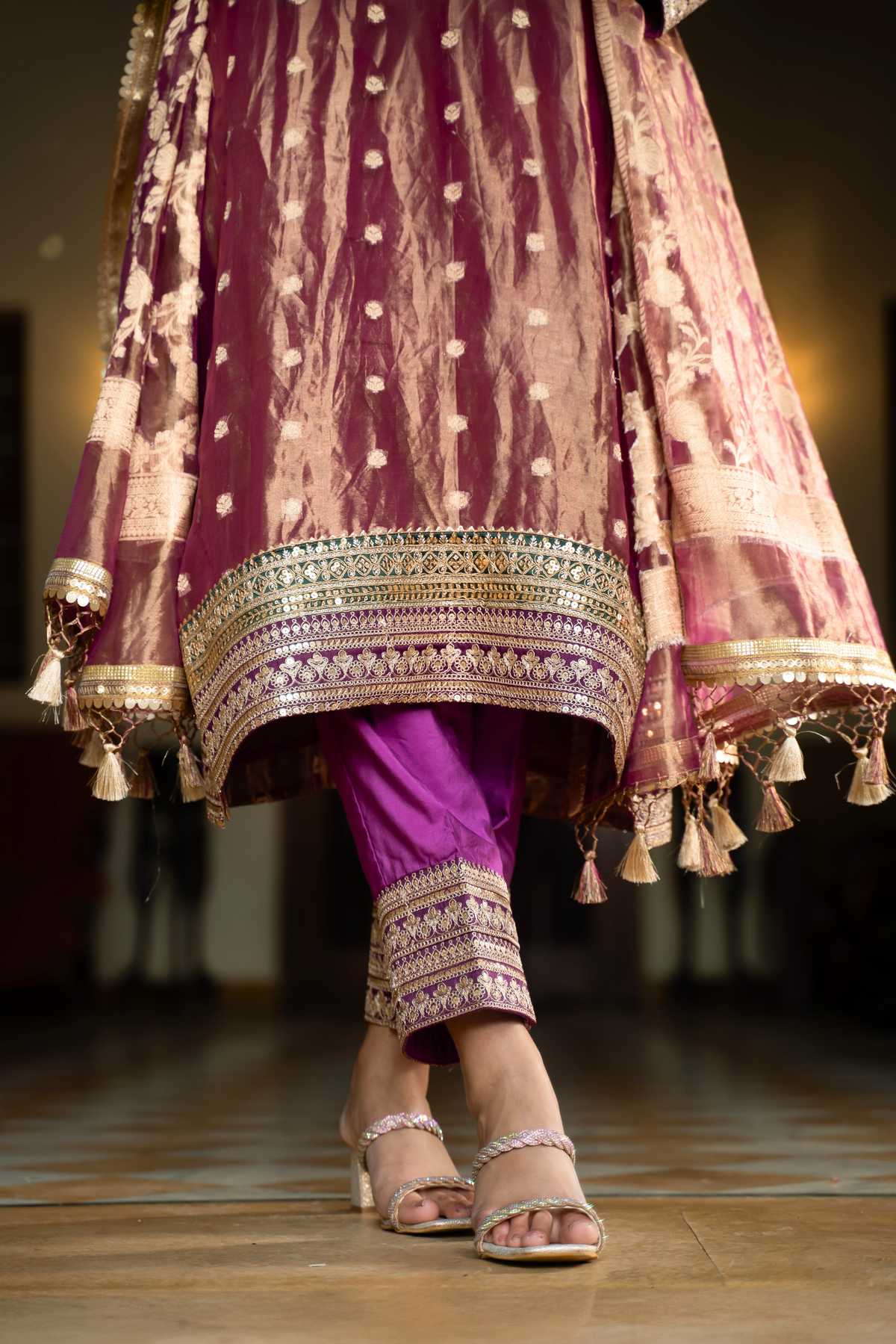 A close-up of a womanβs feet in stylish sandals, wearing a richly embroidered purple and gold traditional banarsi silk kurta set with delicate tassels.