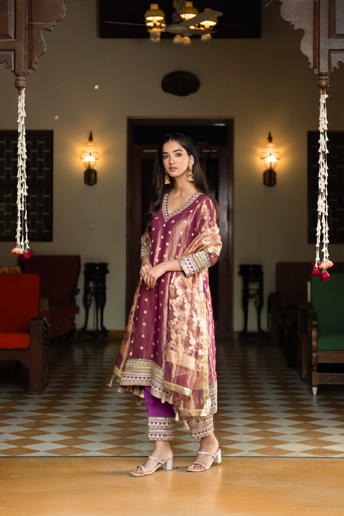 Side view of a woman in a traditional purple and gold anarkali outfit stands elegantly in a vintage-style room, surrounded by decor and plush seating.