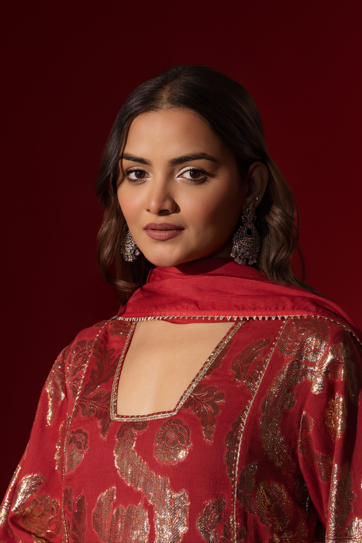 Closeup view of a woman poses in a maroon ethnic shimmer kurta set outfit with intricate white embroidery against a vibrant red backdrop.