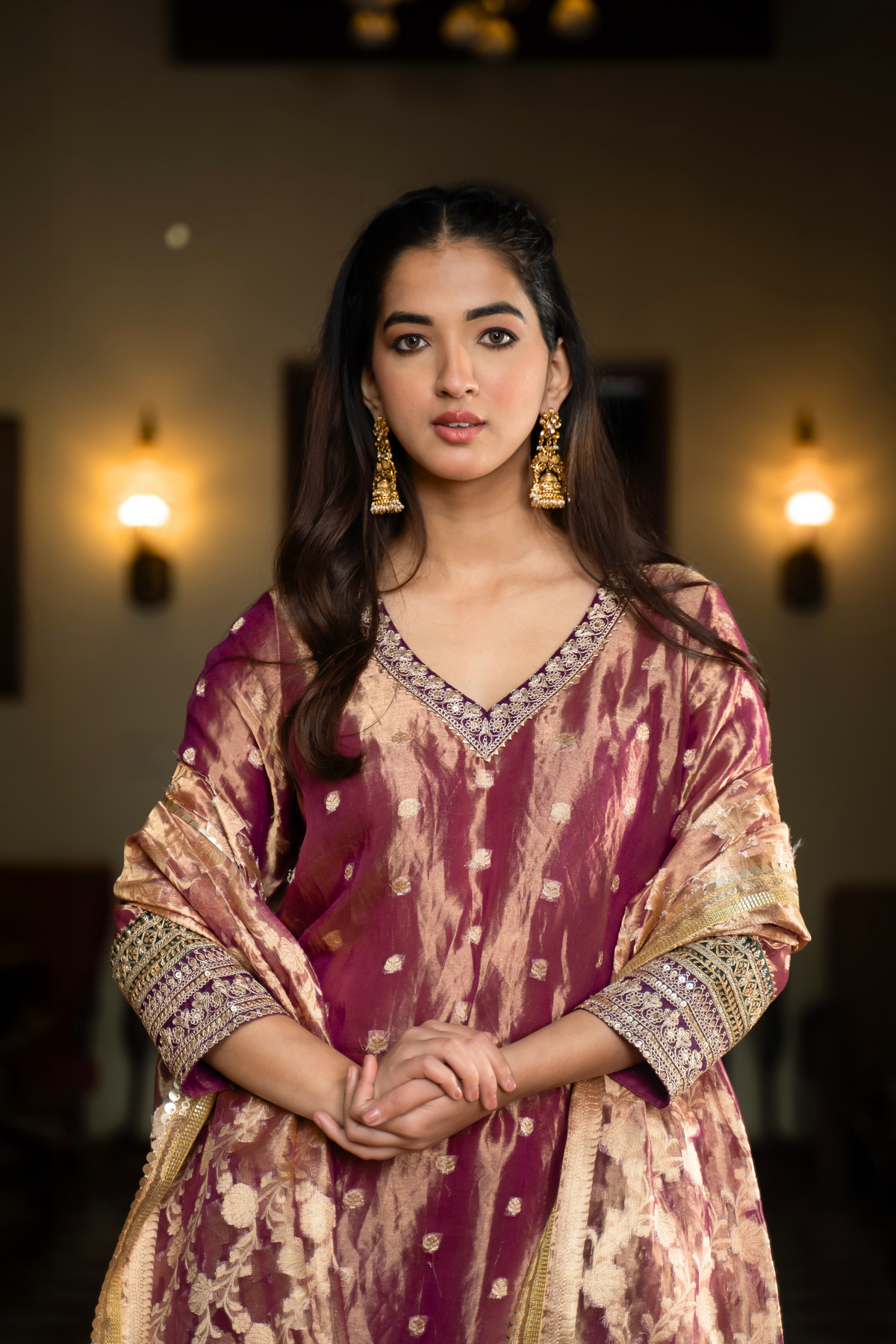 Closeup view of a woman in a traditional purple and gold anarkali outfit stands elegantly in a vintage-style room, surrounded by decor and plush seating.