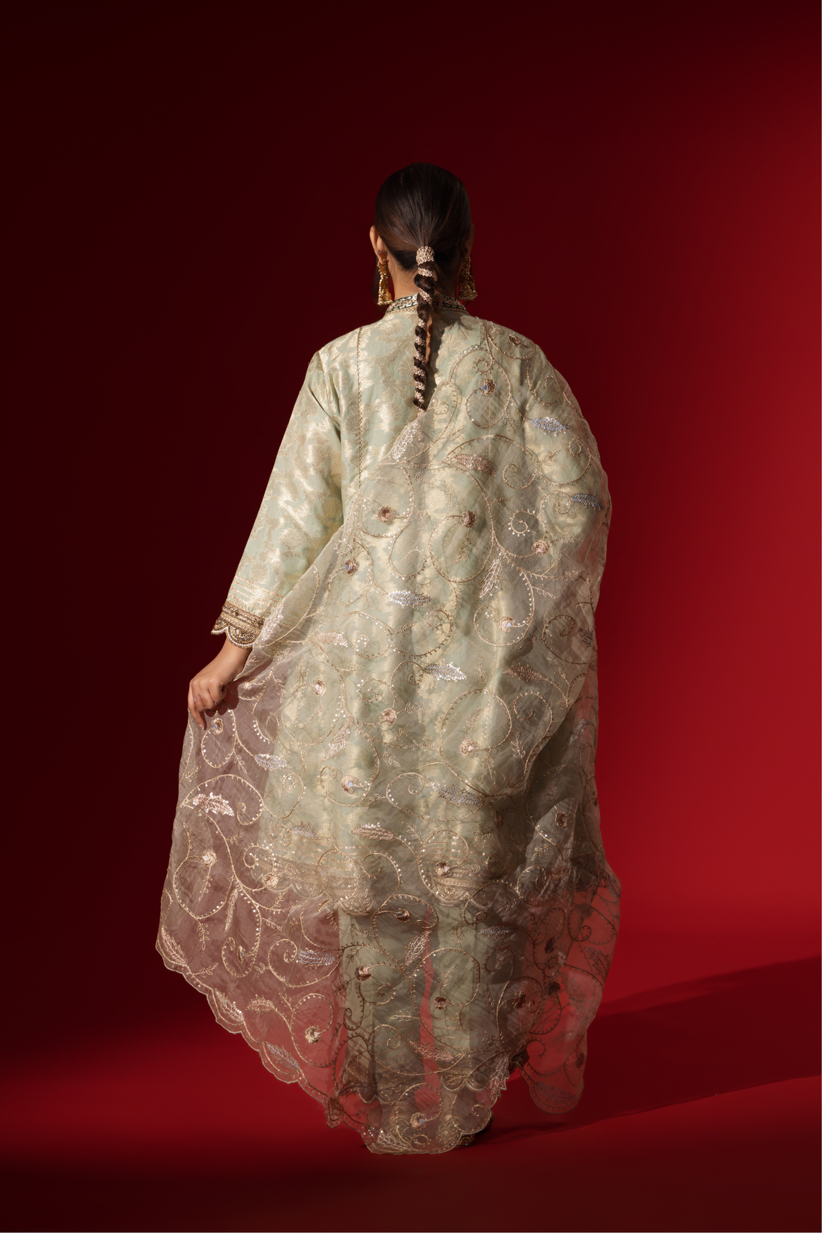 Back view of a woman wearing an elegant mint green embroidered sequin brocade kurta set with gold accents and a sheer dupatta, set against a rich red background
