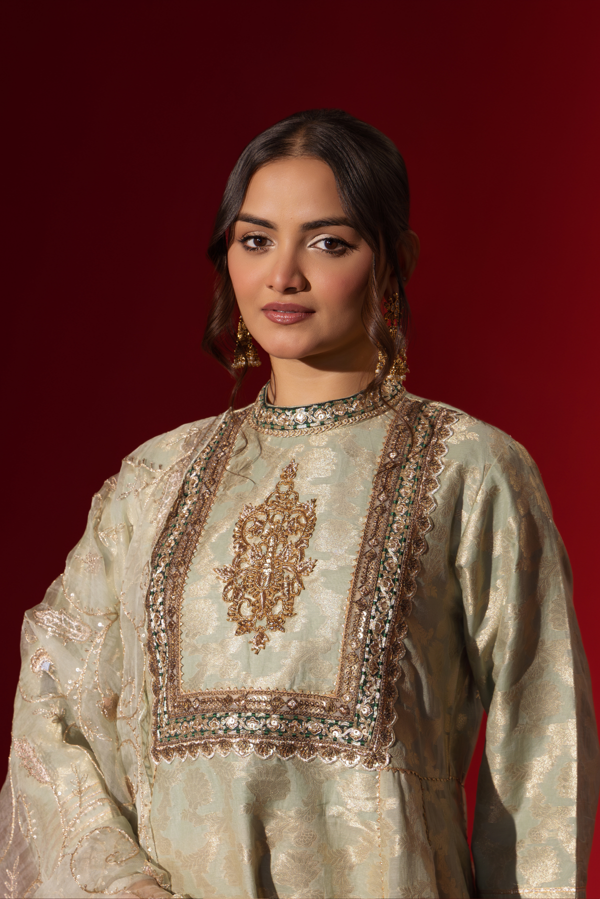 Closeup view of a woman wearing an elegant mint green embroidered sequin brocade kurta set with gold accents and a sheer dupatta, set against a rich red background