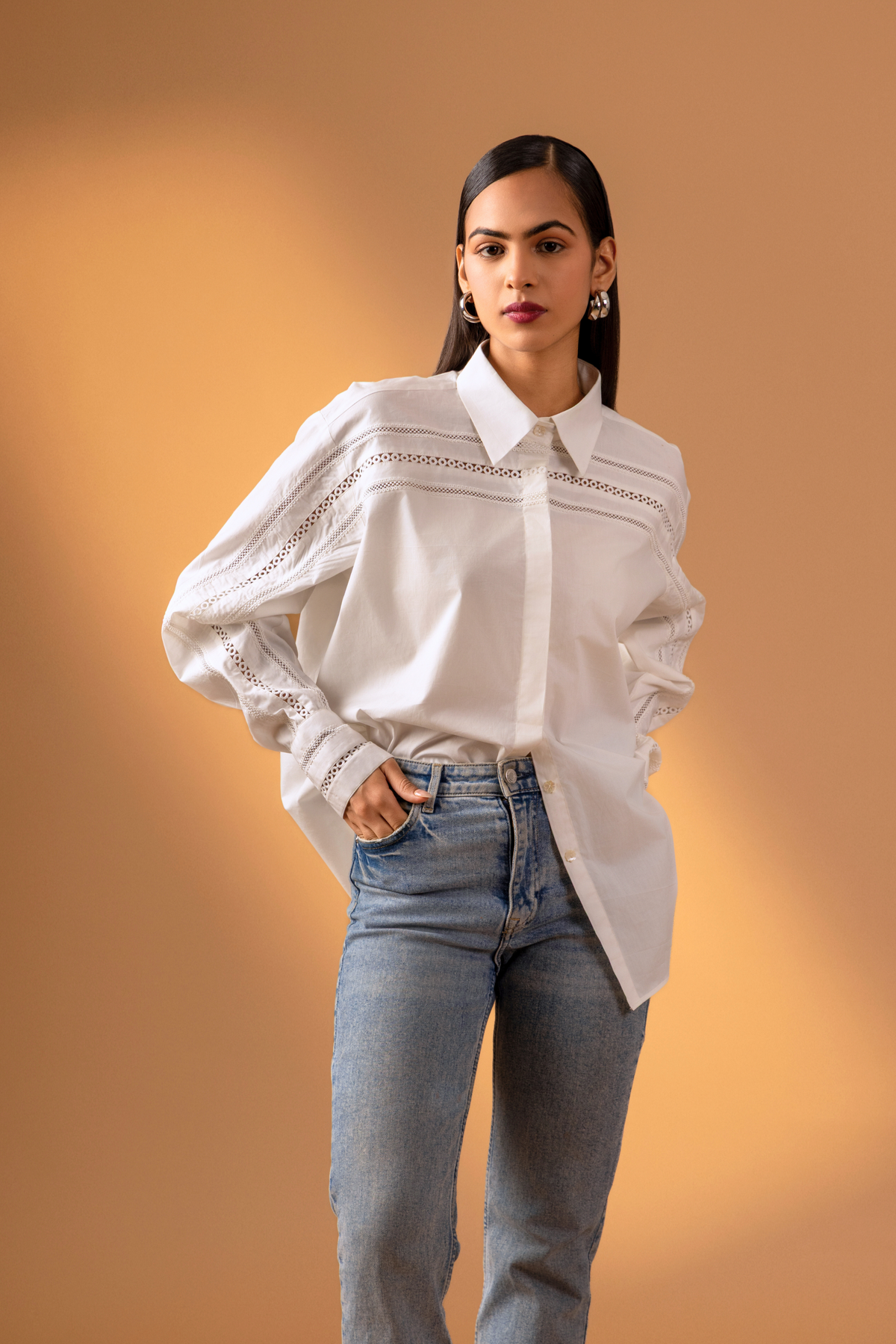 A women stands confidently in a stylish white button-up shirt with intricate details and puffed sleeves, paired with blue jeans & one hand in pocket