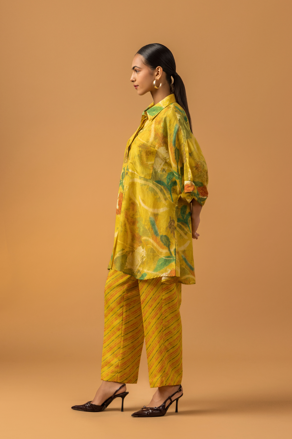 Side view of a model stands in a bright yellow co-ord set featuring a patterned oversized shirt and matching striped pants against a warm backdrop.