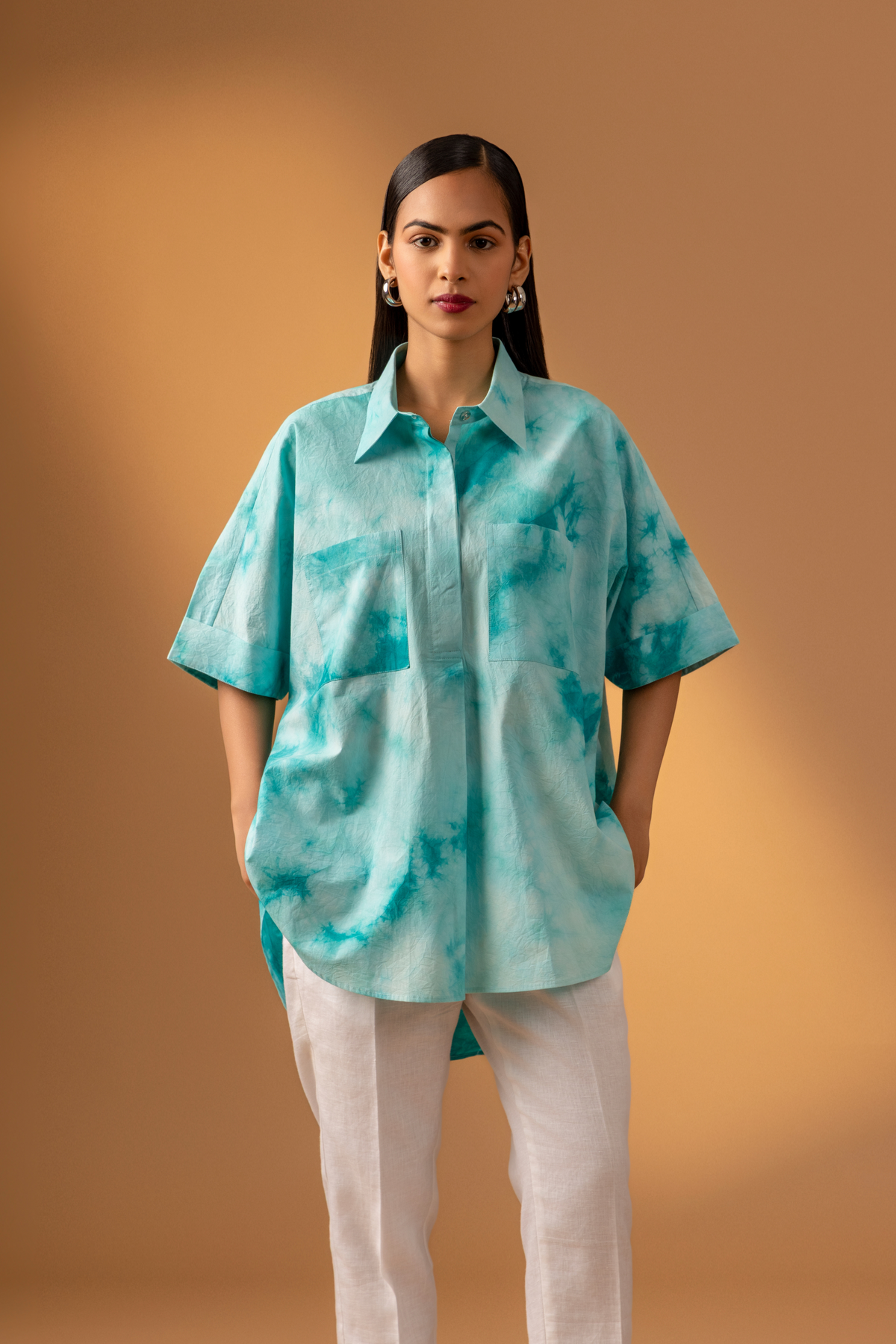 A woman stands confidently in a teal tie-dye oversized shirt and light-colored trousers against a warm, neutral background.