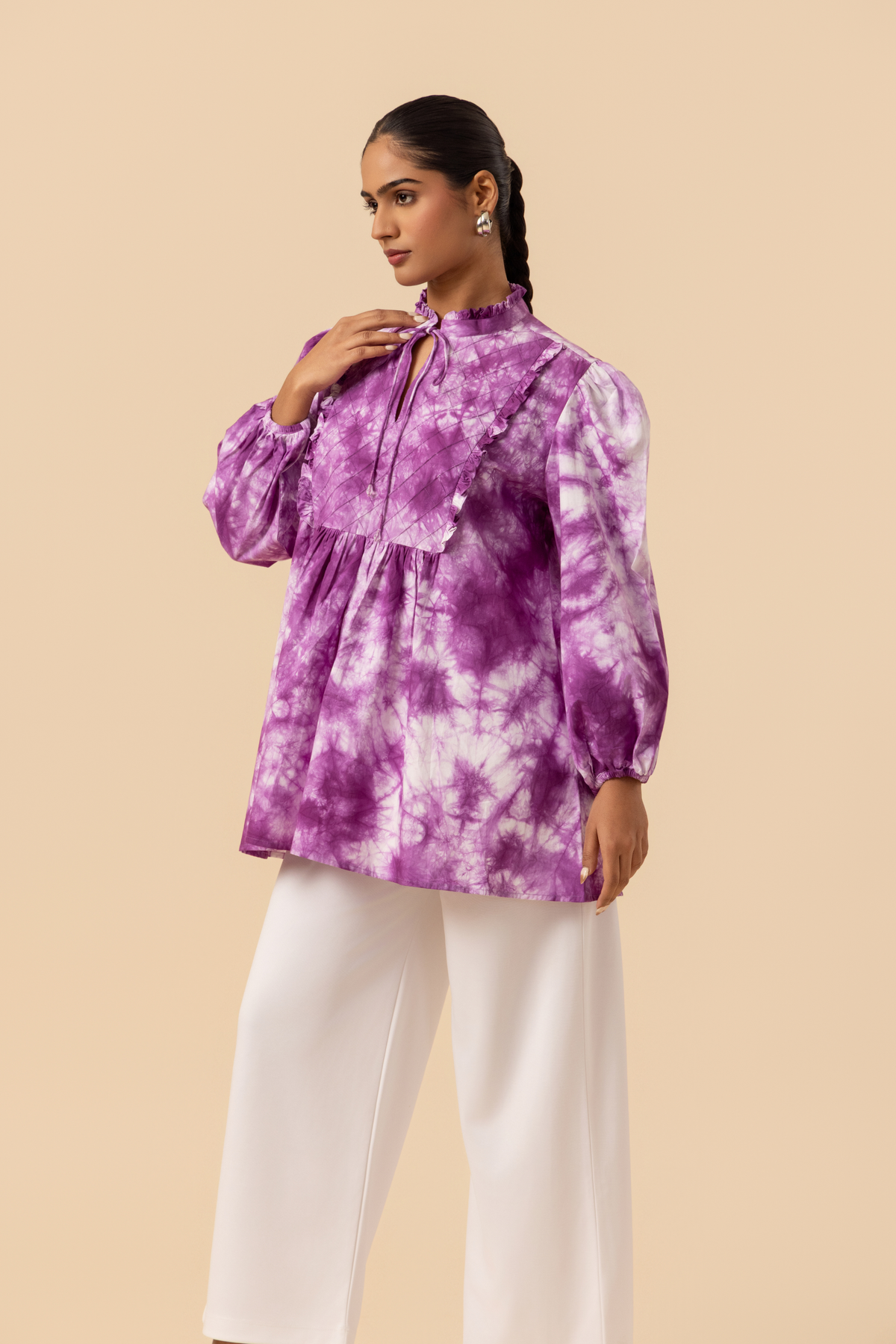 Side view of a model wears a vibrant purple tie-dye shirt dress with puffed sleeves, paired with white wide-leg trousers and stylish sandals.