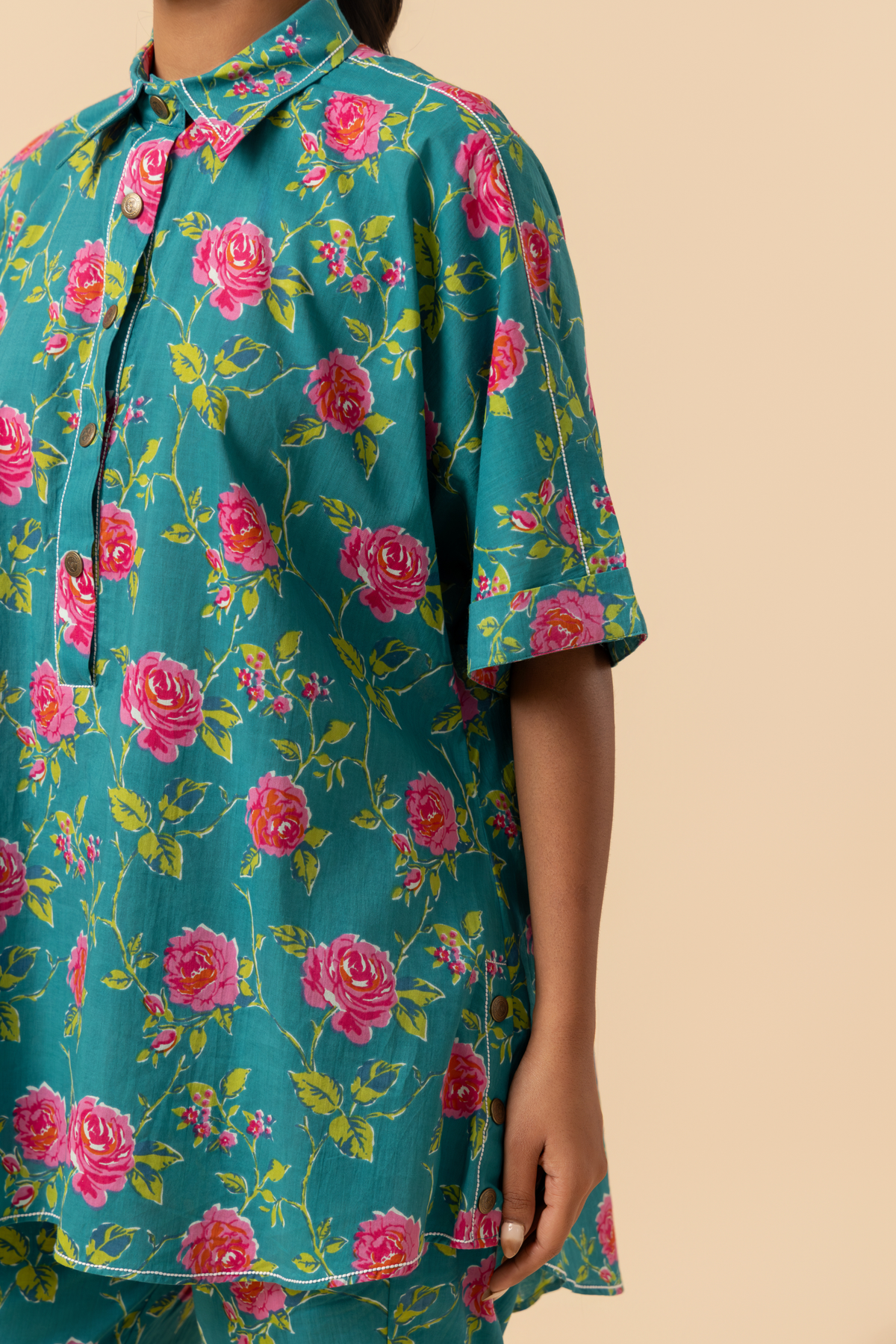 A model stands in a vibrant floral outfit, featuring a teal shirt and matching pants adorned with pink roses, set against a neutral background.