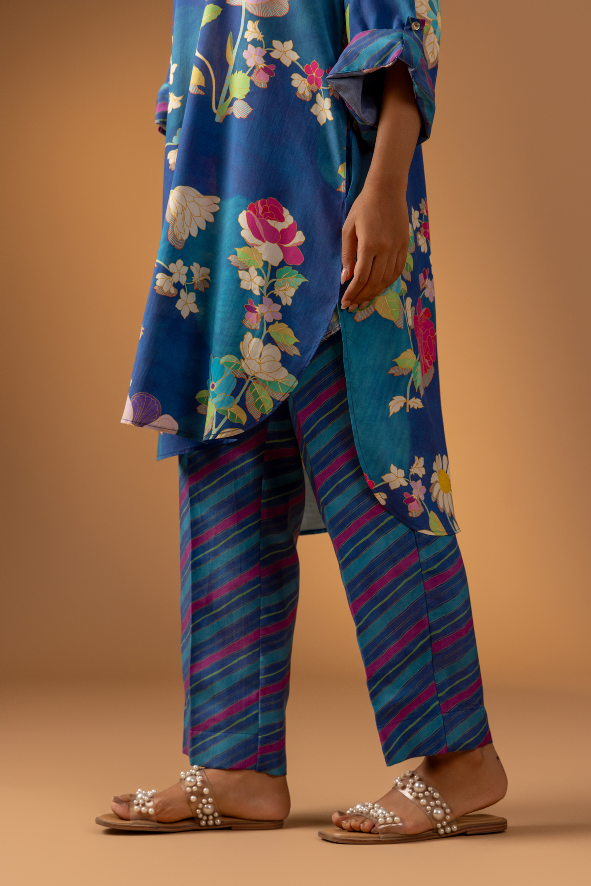 A close-up of a woman woman models a vibrant floral co-ord set paired with striped pants, set against a warm, neutral background.