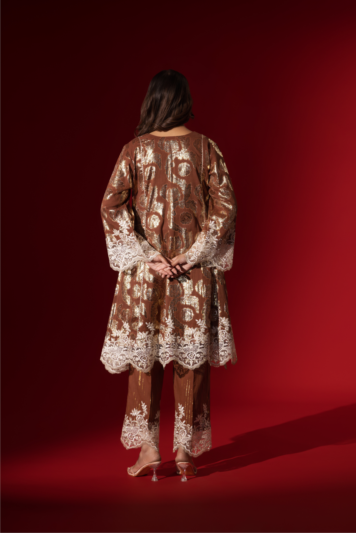Back view of a model poses in a brown ethnic shimmer kurta set outfit with intricate white embroidery against a vibrant red backdrop.