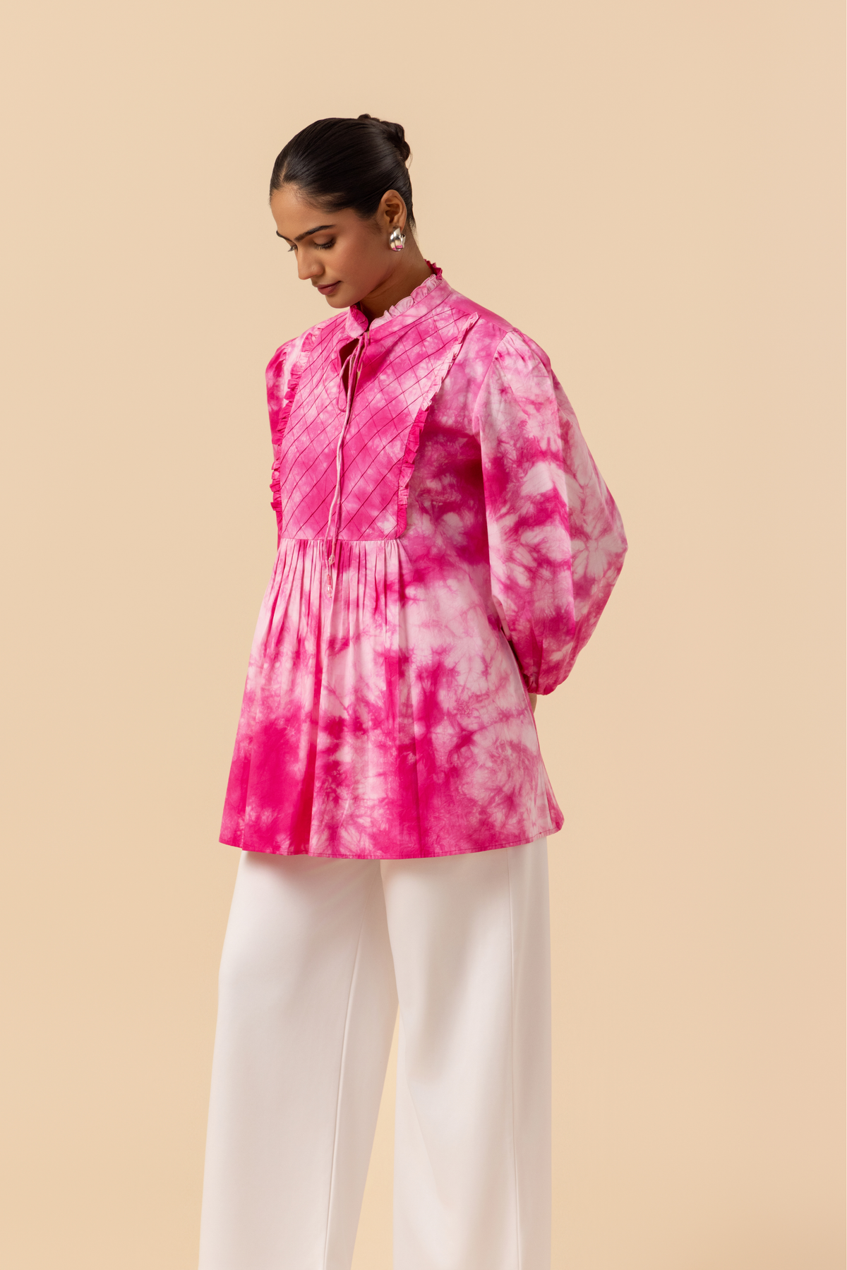 Side view of a model wears a vibrant pink tie-dye shirt dress with puffed sleeves, paired with white wide-leg trousers and stylish sandals.