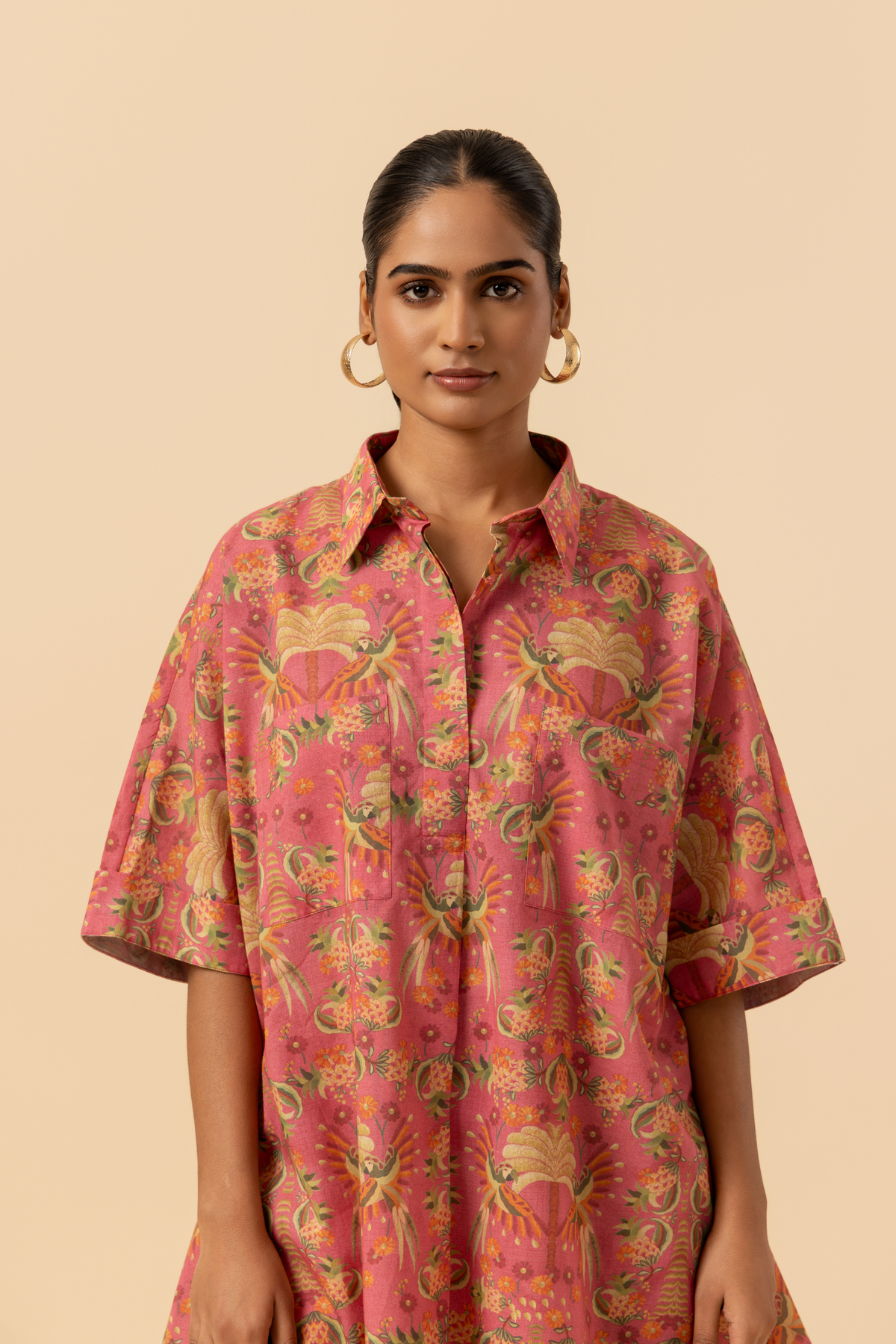 Closeup view of a woman stands in a vibrant pink and floral-patterned co-ord set, featuring a loose shirt and matching pants, set against a neutral background.