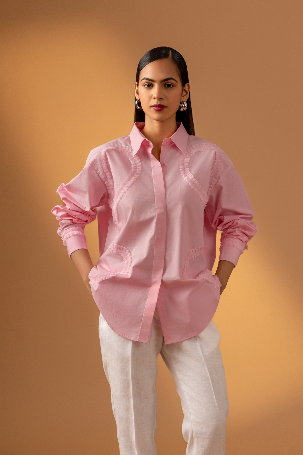 A woman stands against a warm, neutral backdrop, wearing a pink oversized shirt, white trousers, and stylish sandals & hands in pockets.