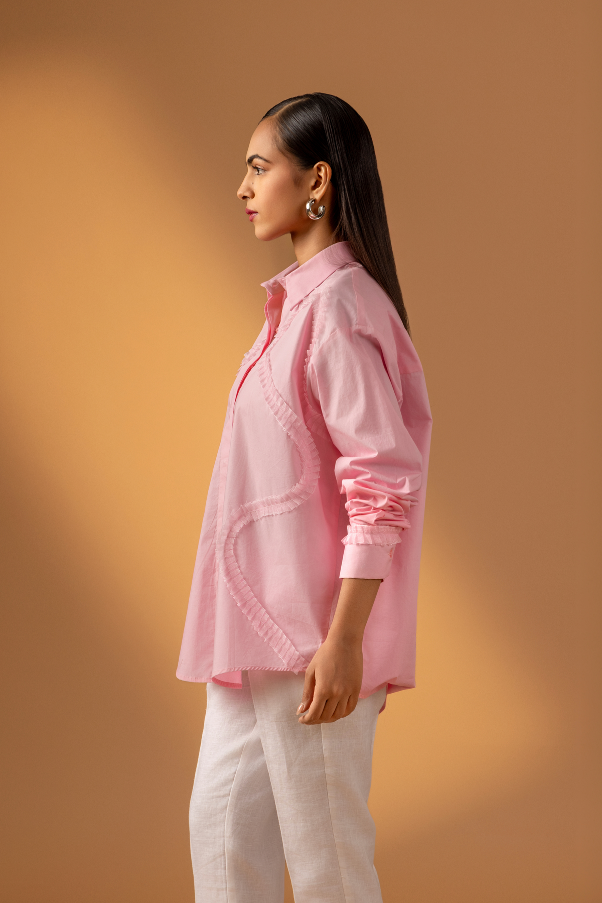Side view of a woman stands against a warm, neutral backdrop, wearing a pink oversized shirt, white trousers, and stylish sandals.