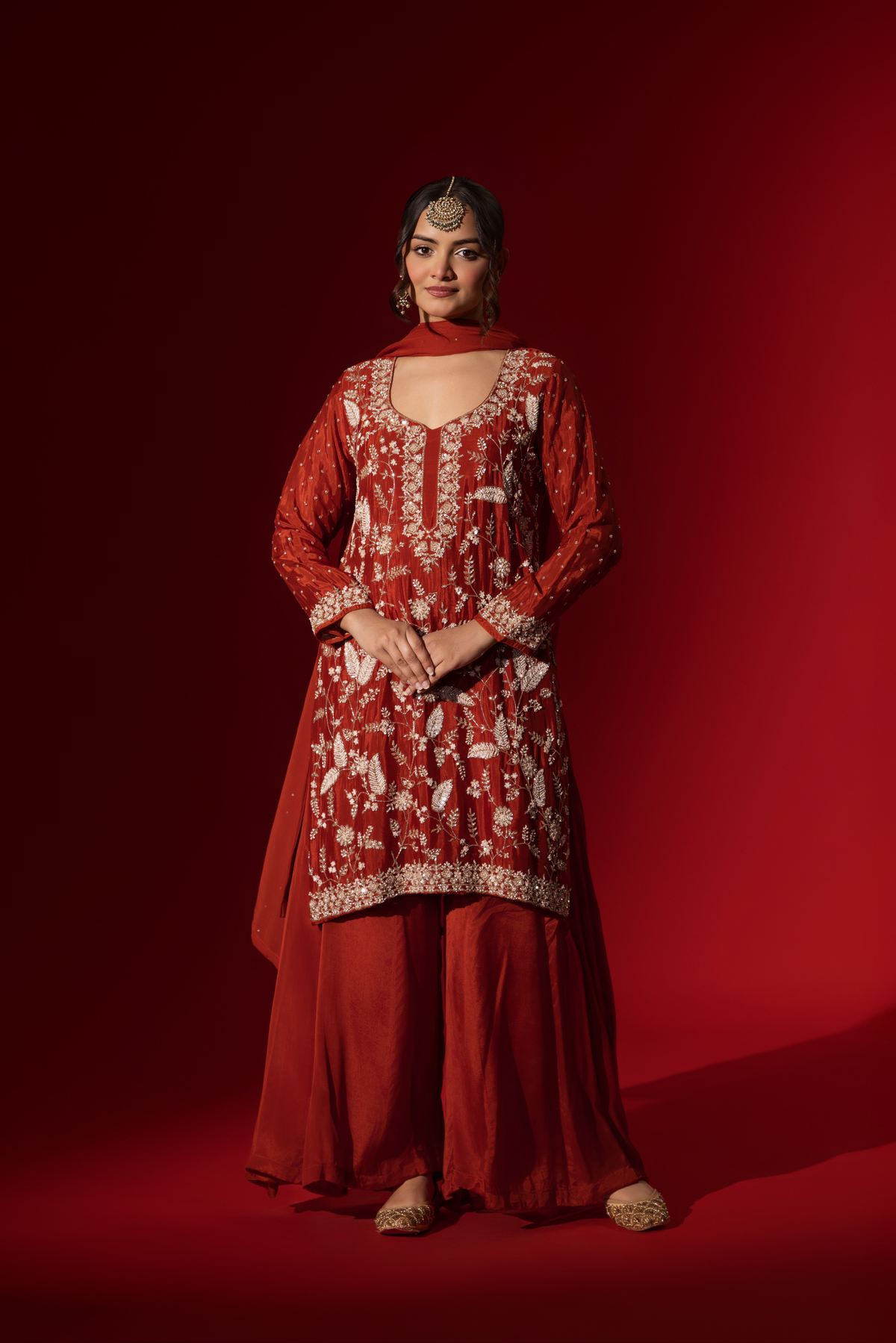 A woman poses in an elegant red embroidered kurta set adorned with intricate white embroidery against a rich red background.