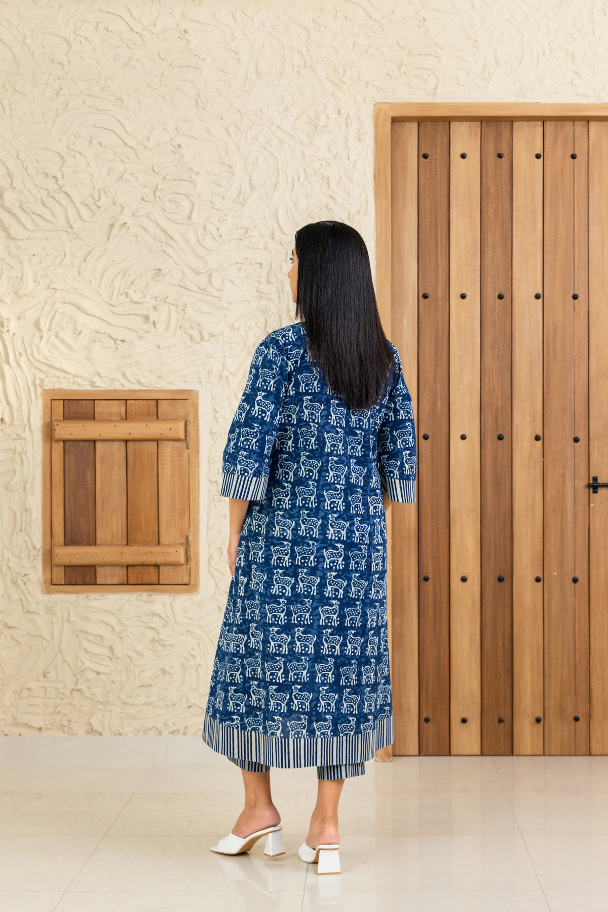 Back view of a woman wearing a blue and white deer printed co-ord set with striped detailing, standing against a wooden and textured beige background.
