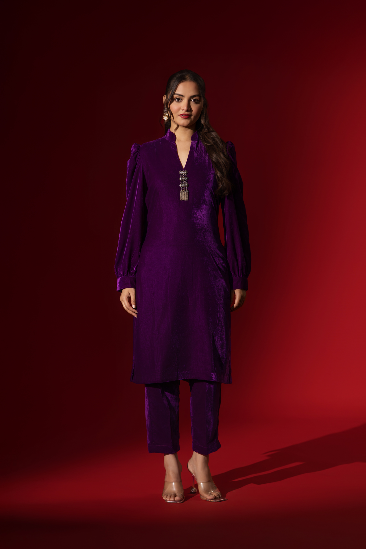 A woman in a vibrant purple velvet co-ord set with a feminine silhouette and elegant earrings, set against a rich red background.