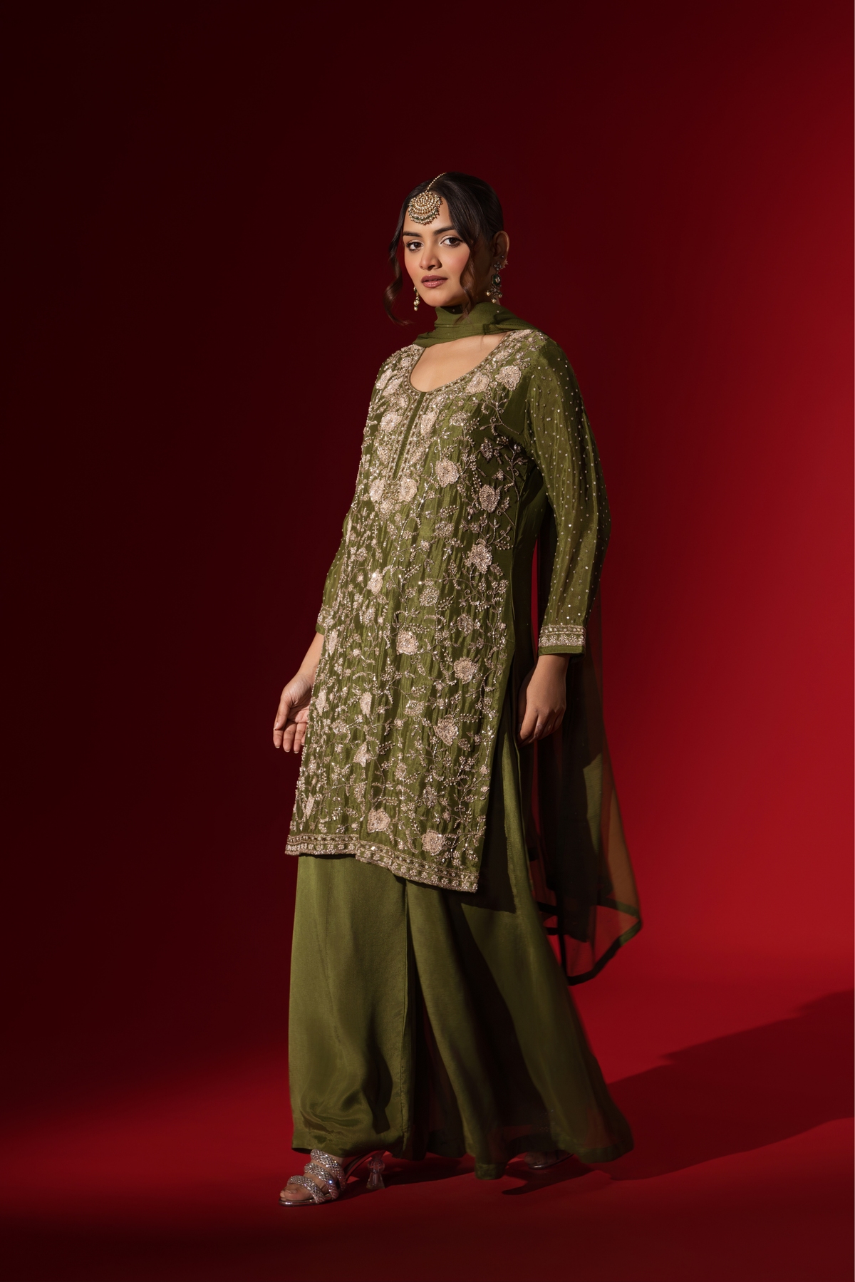 Side view of a woman poses in an elegant olive green embroidered kurta set with intricate floral embroidery, set against a rich red backdrop.