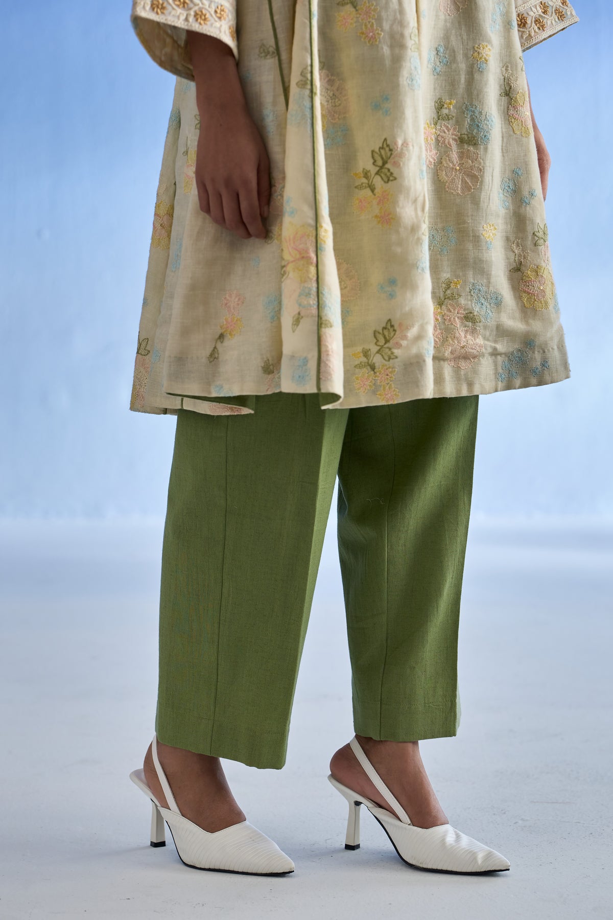 A model is shown from the waist down wearing a beige embroidered linen kurta set, olive green cropped trousers, and white slingback kitten heels against a light blue studio background.
