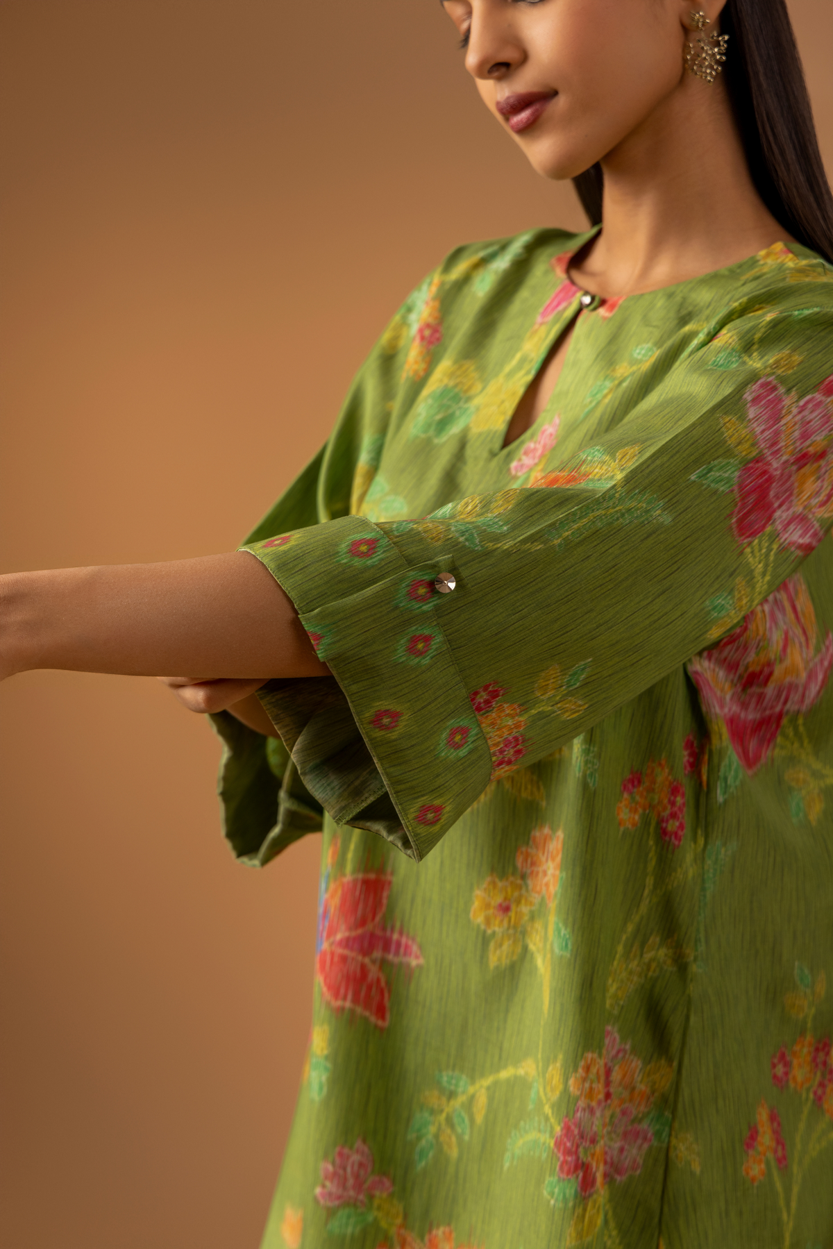 Closeup view of a model showing her hand in a vibrant green co-ord set with floral patterns, paired with matching trousers, against a warm beige background.