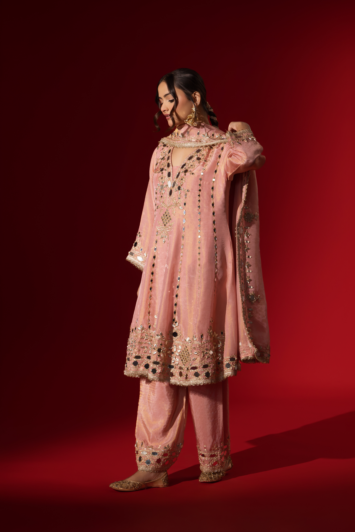 Side view of a person stands against a red background, wearing an elegant pink embroidered kurta set intricately embellished outfit with a matching shawl.