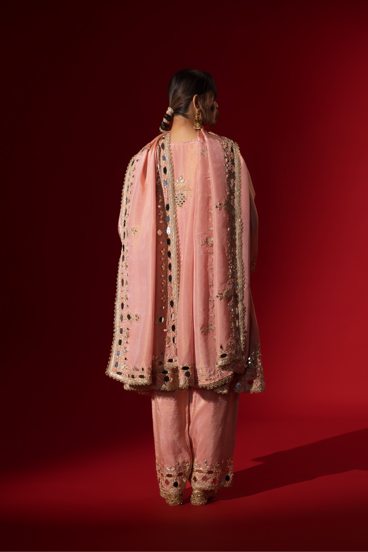 Back view of a person stands against a red background, wearing an elegant pink embroidered kurta set intricately embellished outfit with a matching shawl.