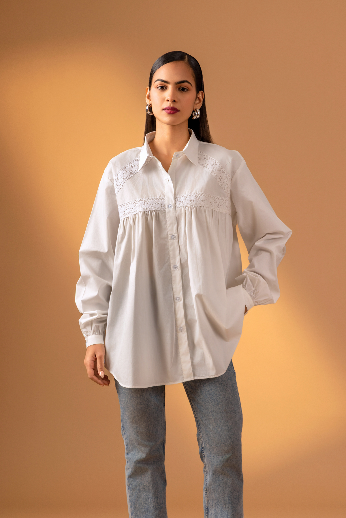 A model wearing a white button-down shirt with lace detailing, paired with blue jeans, & one hand in pocket set against a warm beige backdrop.