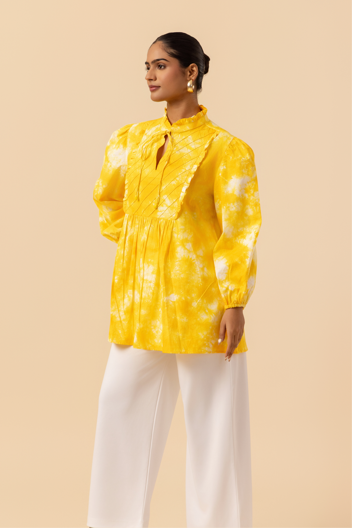 Left side view of a woman wears a vibrant yellow tie-dye shirt dress with puffed sleeves, paired with white wide-leg trousers and stylish sandals.